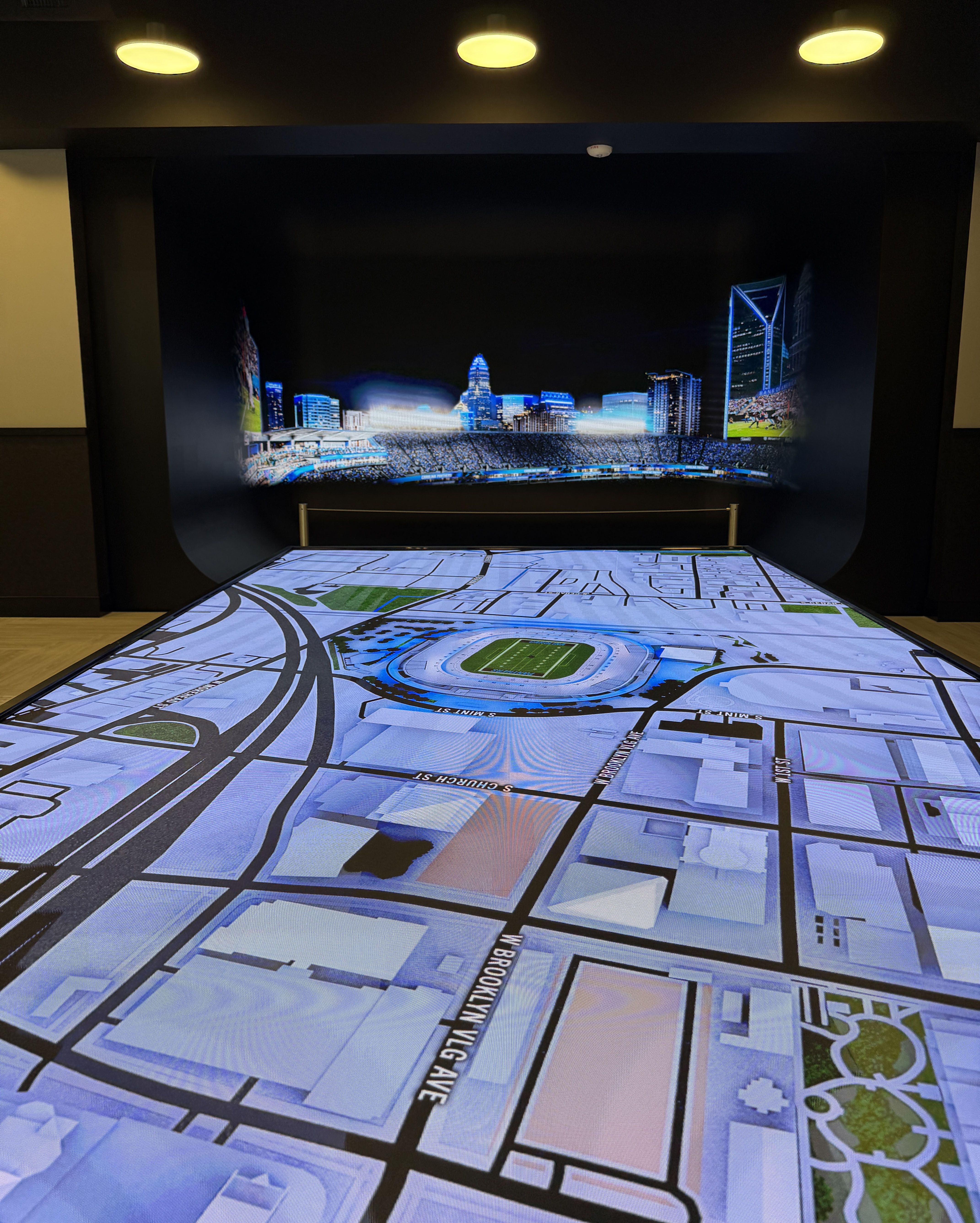 An indoor exhibit features a large printed floor map with streets and a football stadium, set before a blue-lit city skyline mural; three yellow ceiling lights illuminate a dark wall.