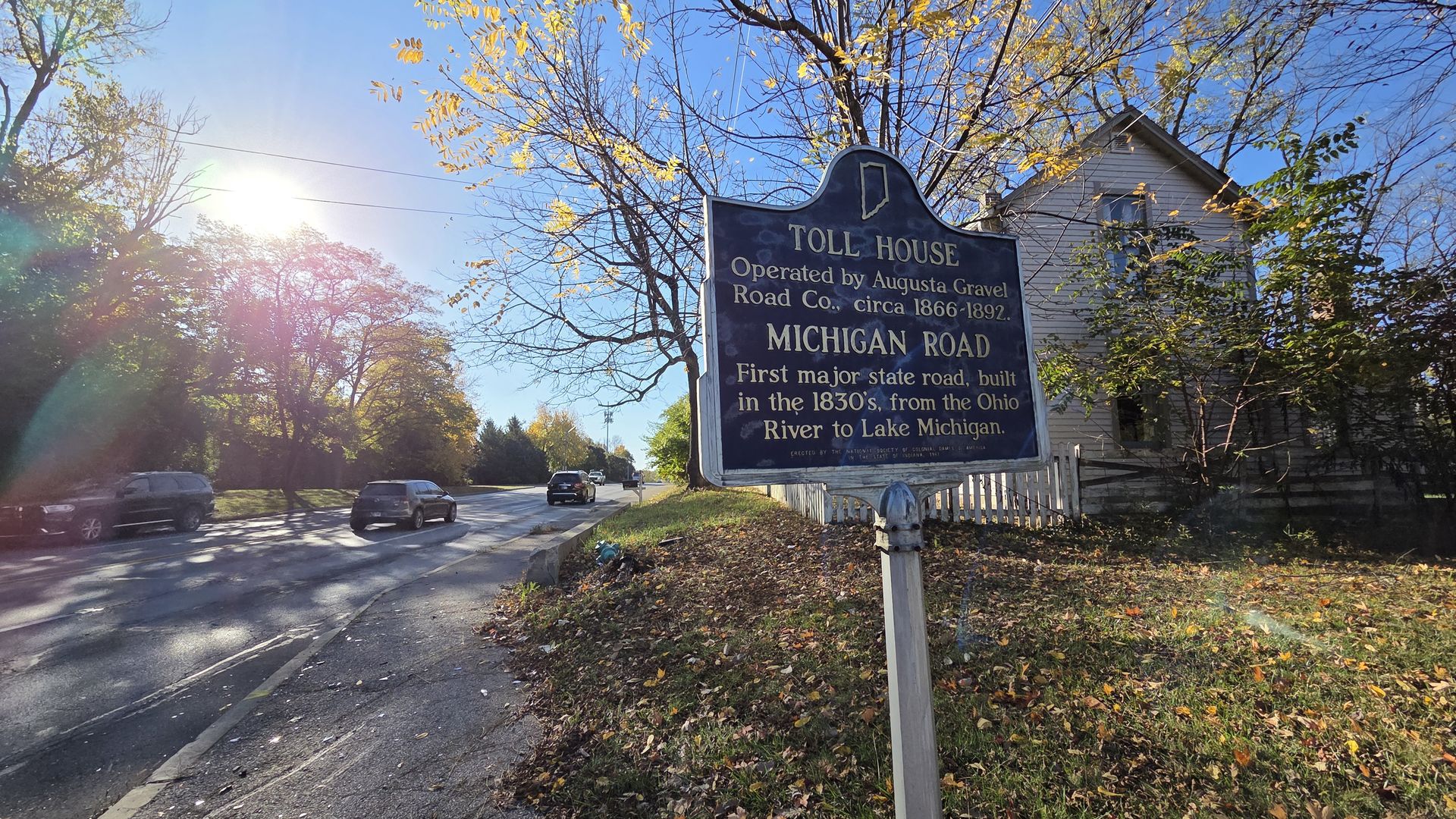 Blue historical sign about Toll House and Michigan Road by a sidewalk with autumn leaves, trees, cars on road, and a white house under a clear blue sky with sun glare.