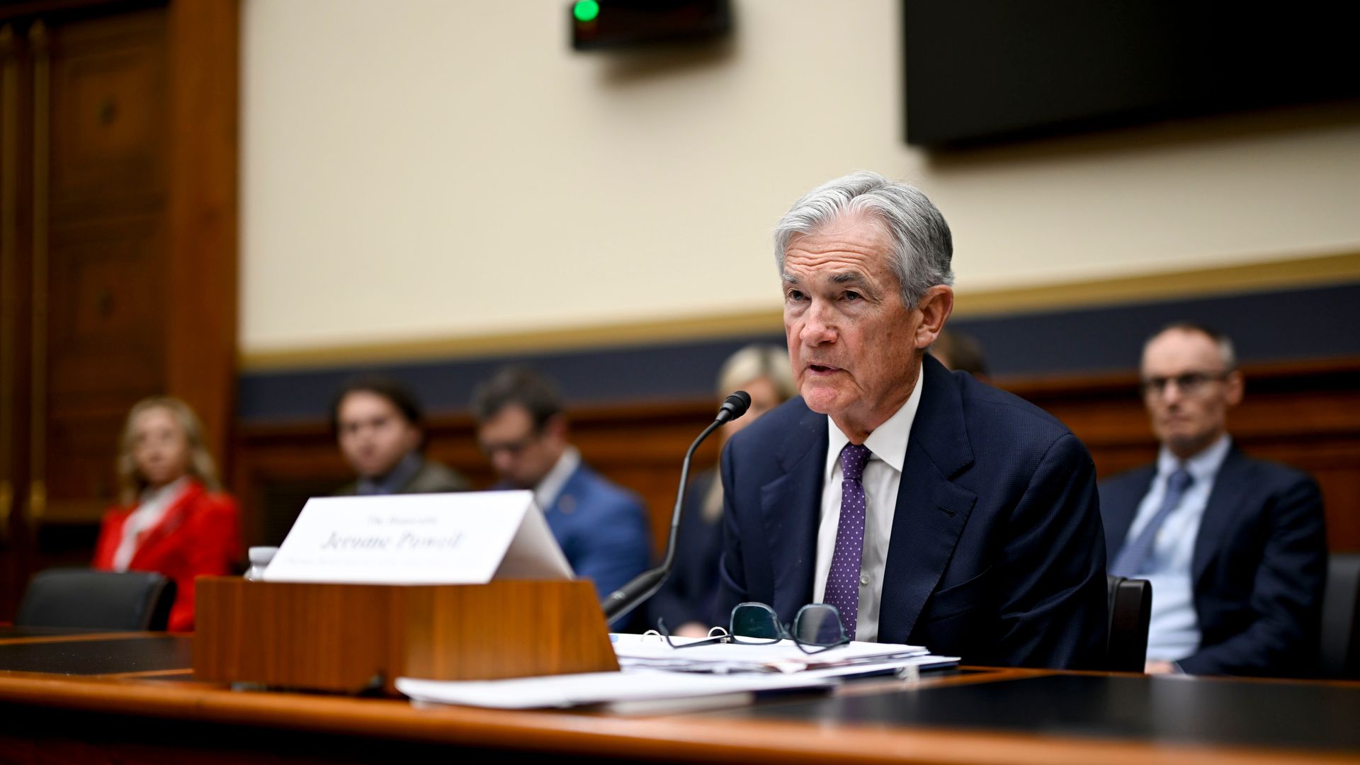 A man sitting at a desk speaking with papers in front of him, his glasses on the table. Blurry people are sitting behind him. 
