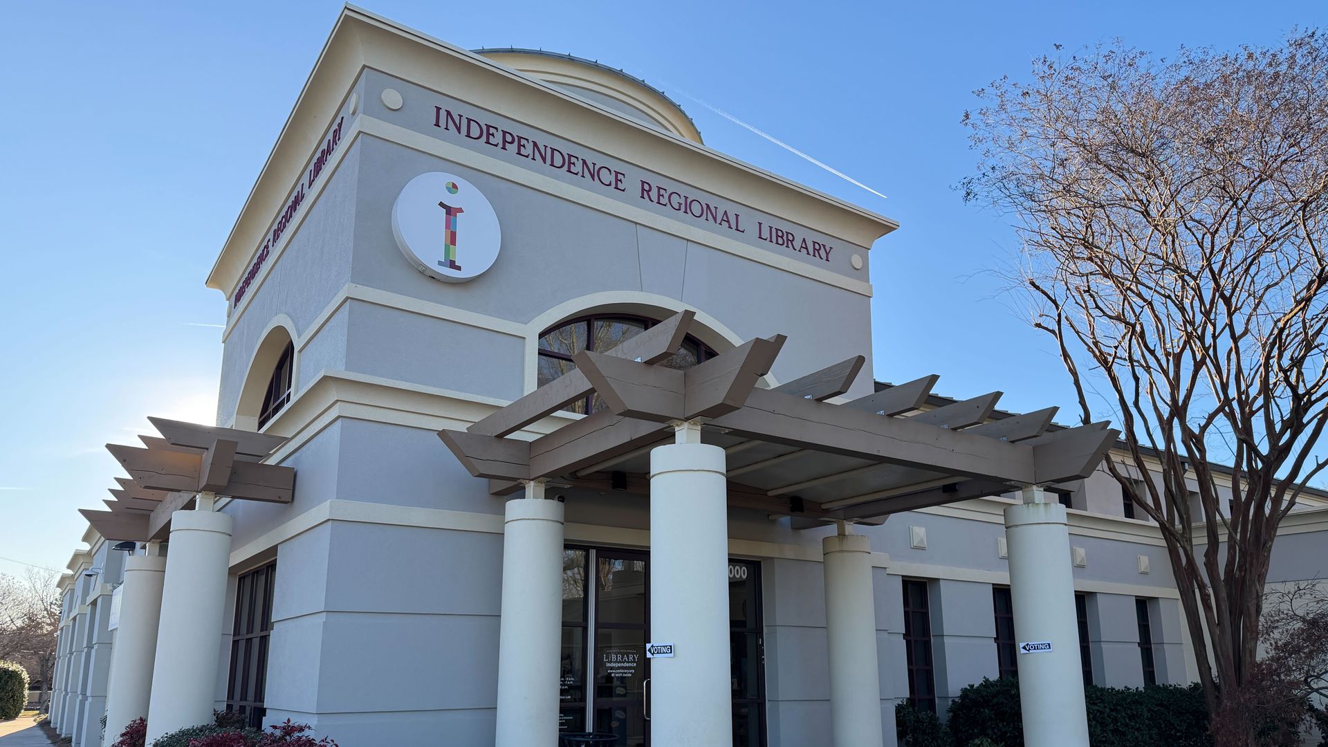 Exterior of the Independence Regional Library building with beige columns, a wooden pergola, purple window frames, and leafless trees under a clear blue sky.