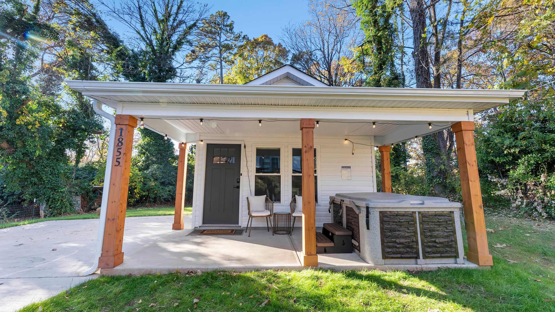 Small white house with a covered porch supported by wooden beams, two chairs with cushions, a side table, and a gray hot tub, surrounded by green grass and tall trees under blue sky.