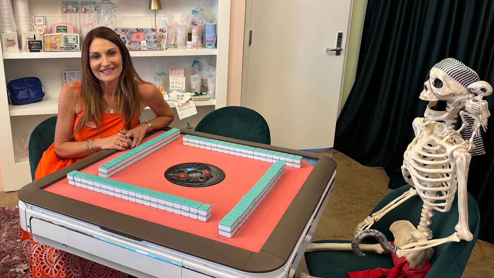 Smiling woman in an orange dress sits at a pink mahjong table with turquoise tiles; white shelves behind display gifts and mugs. To the right, a striped-cap skeleton sits near a door labeled "Storage Closet".
