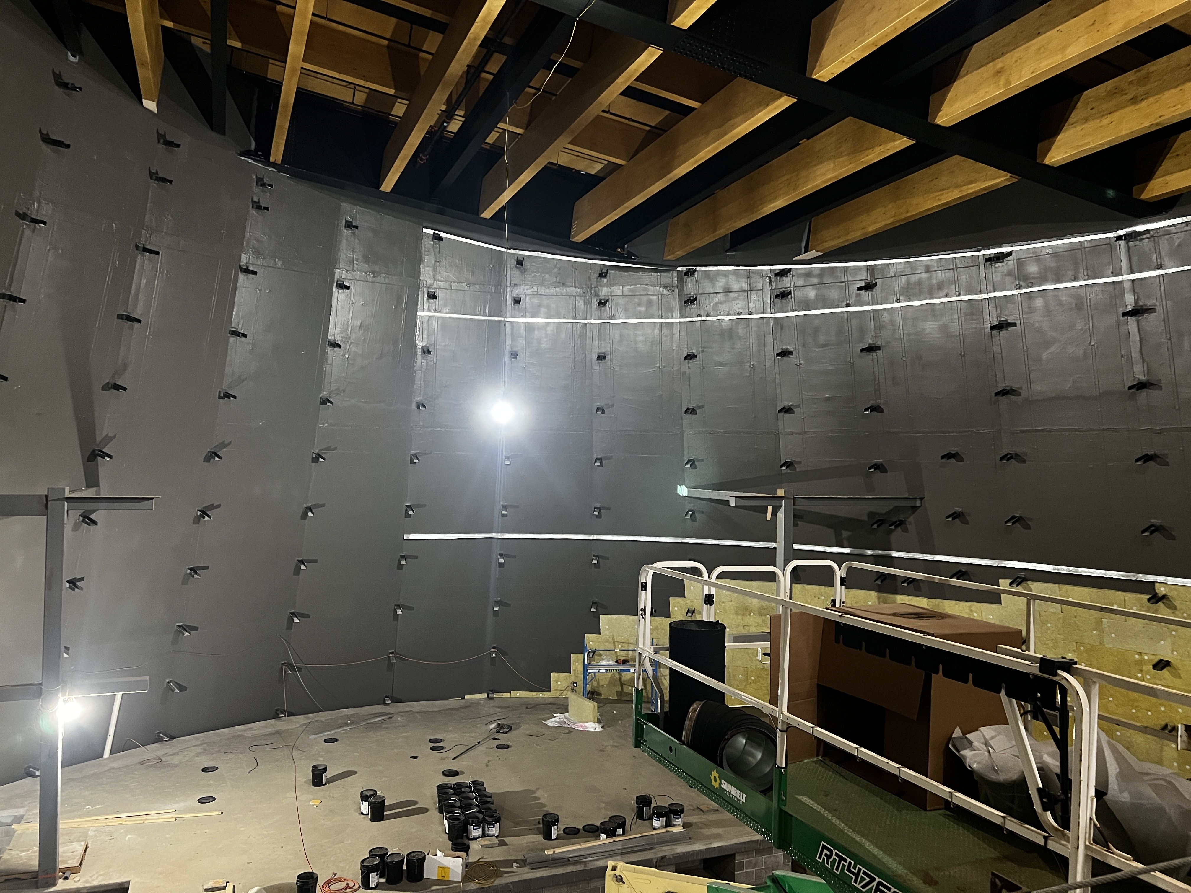 Indoor construction scene: curved gray wall with many black fasteners, exposed wooden ceiling beams, bright work lights, and a green scissor lift on the right; tools and cables on the floor.