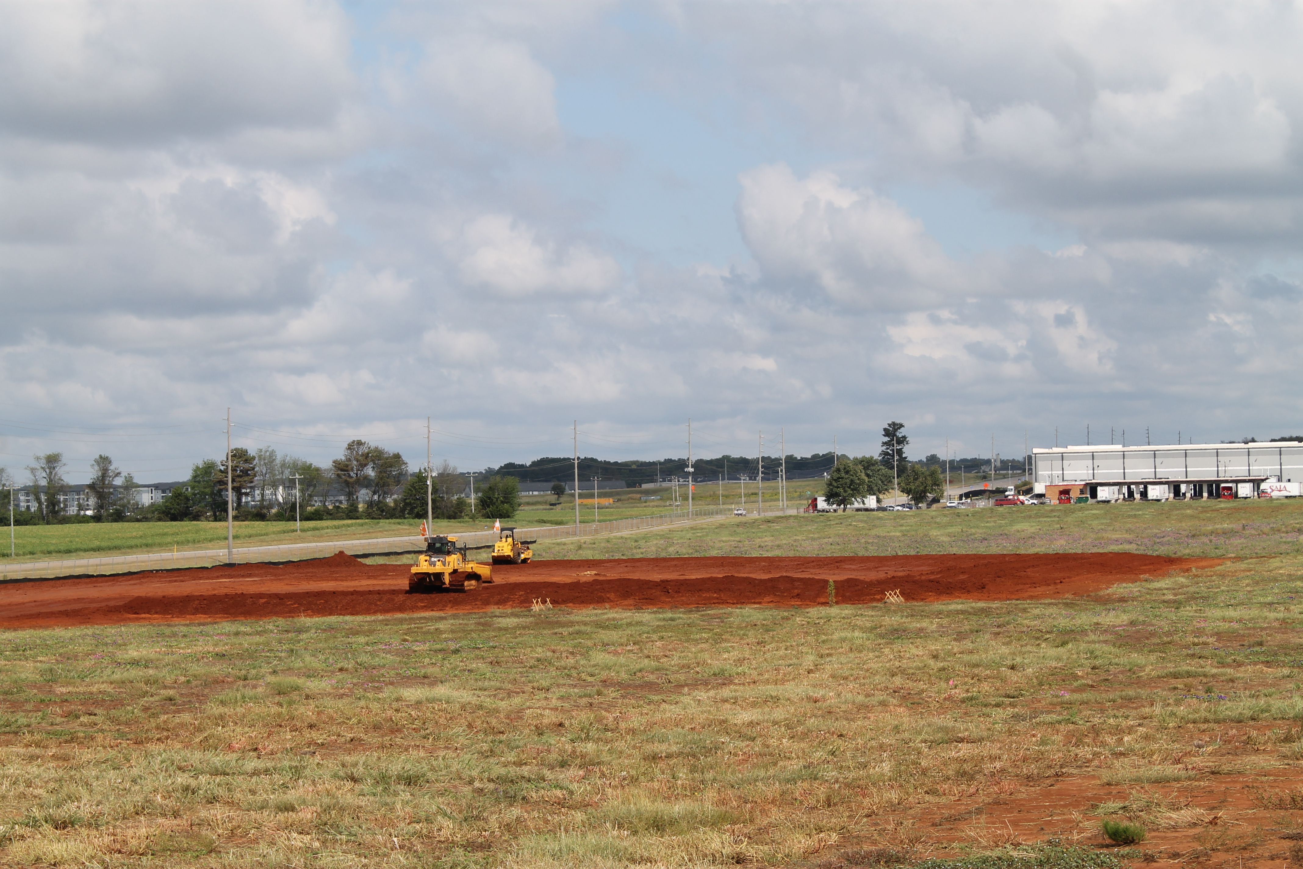 Wide daytime construction site with red earth in the foreground, two yellow road rollers, grassy fields, utility poles and trees, and a long white building with trucks beside a cloudy blue sky.