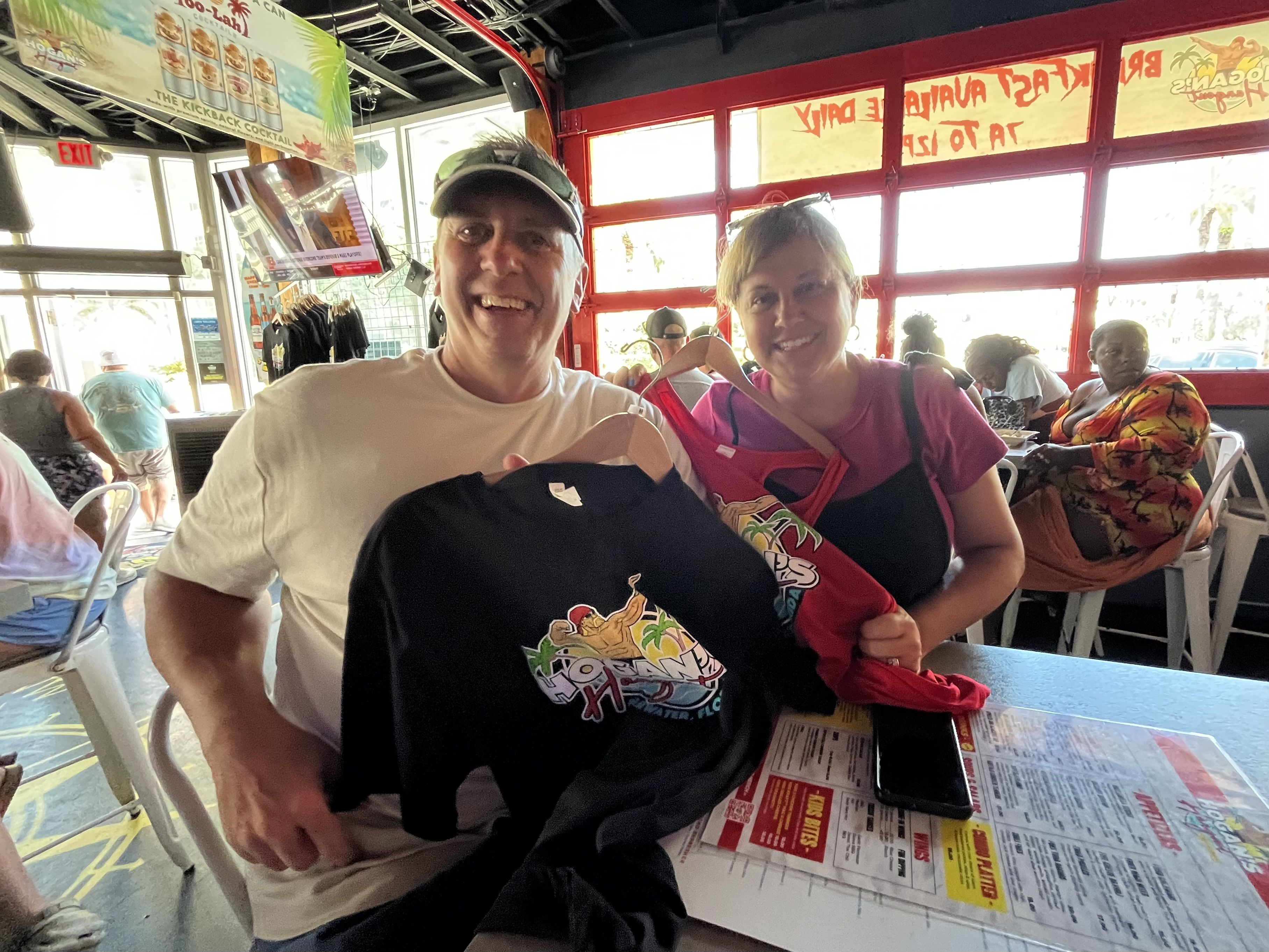 Smiling man in white shirt and visor and woman in pink shirt at a table holding black and red shirts with "Hogans" tropical logo inside a lively, colorful restaurant.
