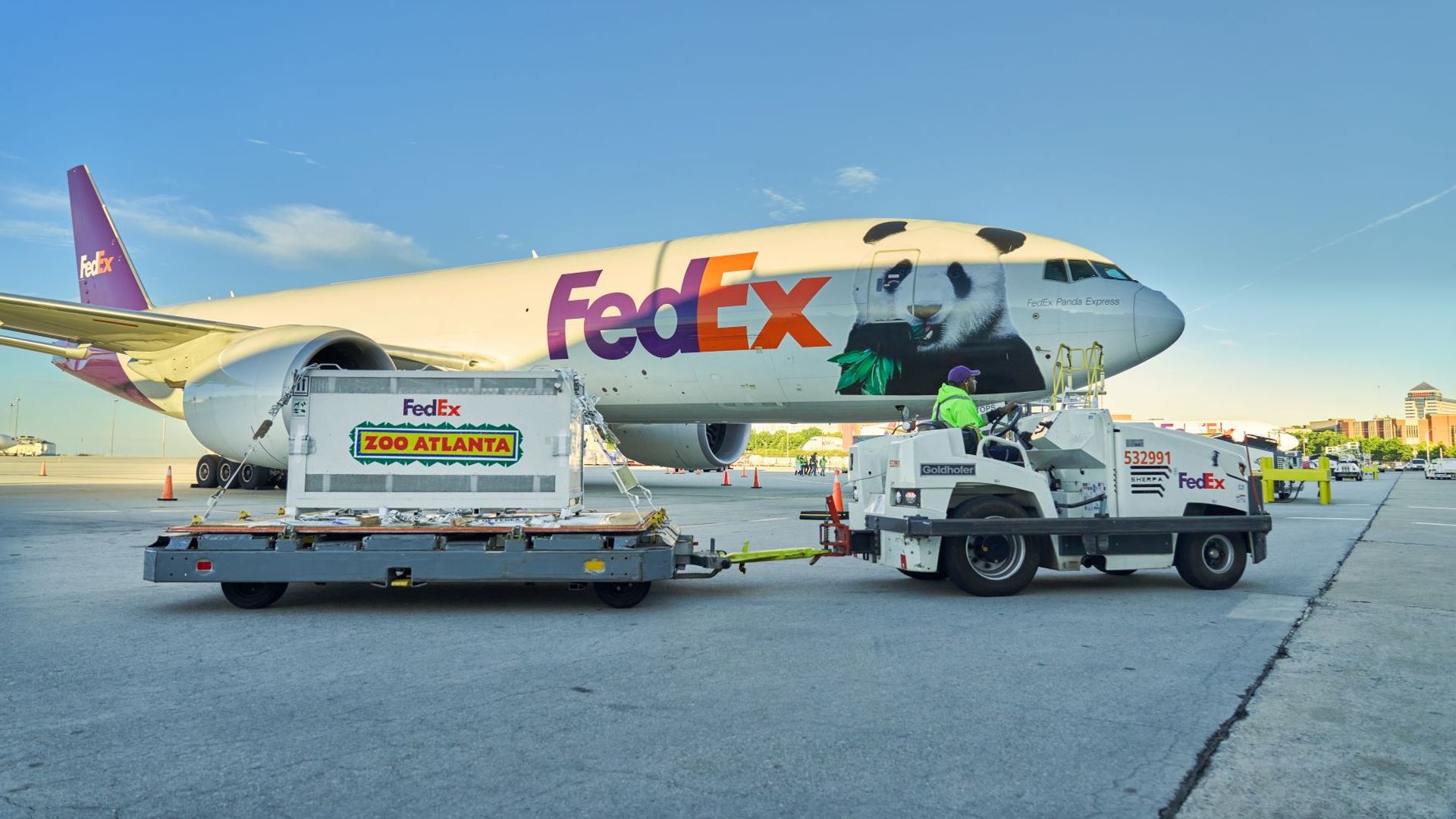 A FedEx cargo plane on the tarmac with a panda graphic on its white fuselage; a loading platform displays a crate labeled "Zoo Atlanta" as ground crew works nearby beneath a clear blue sky.