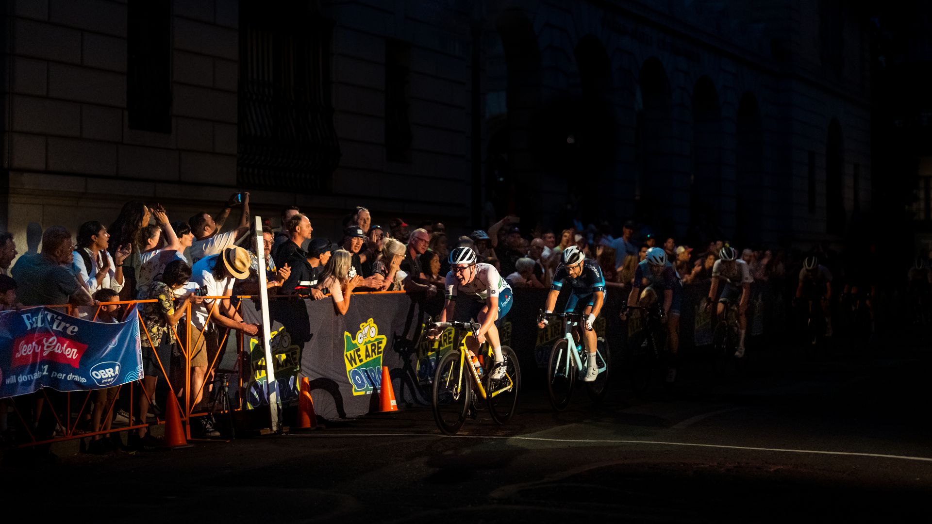 A photo of a bicylist grinning while a crowd of people behind a barricade cheers on.