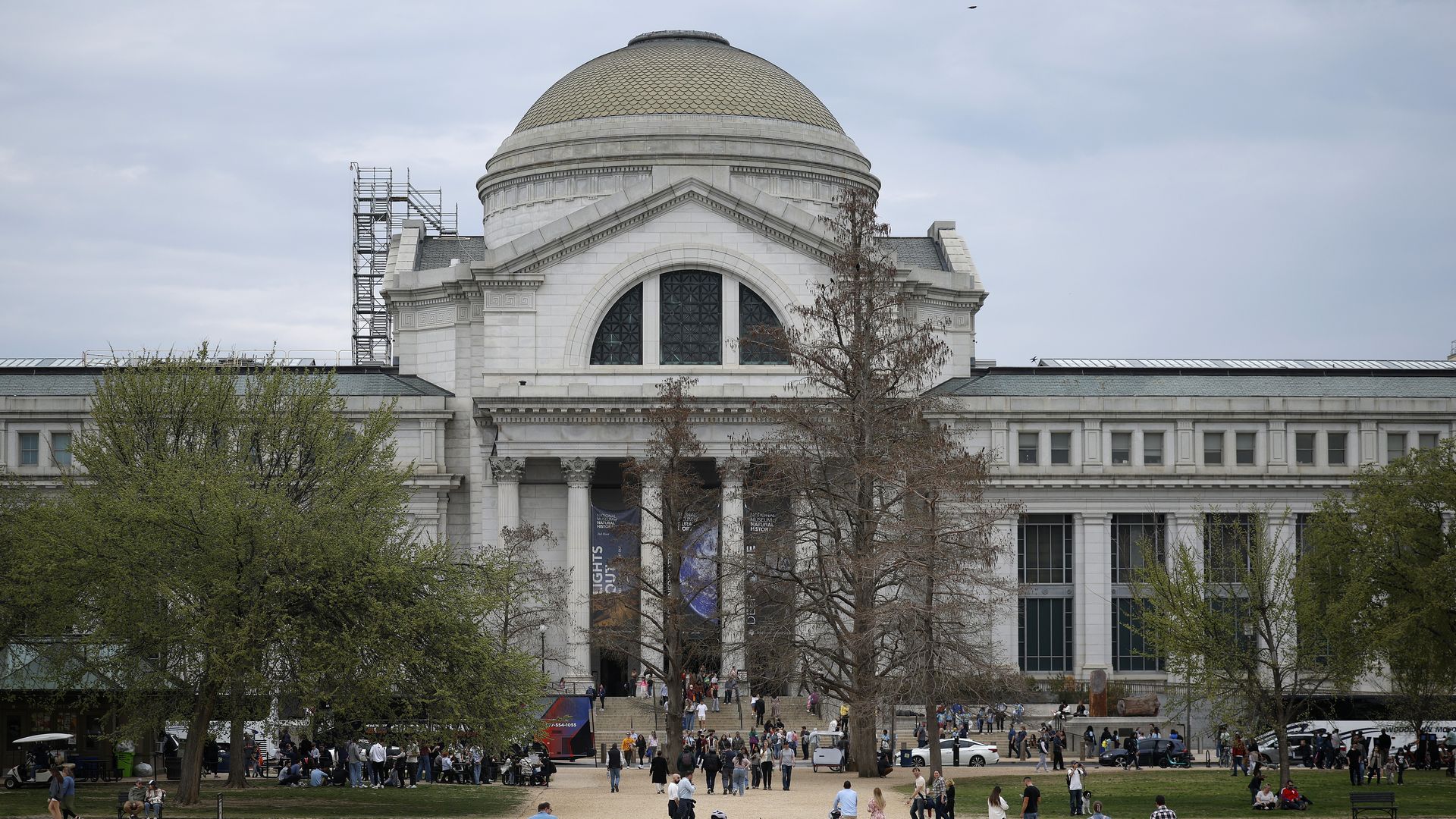 Crowds of people walking and sitting on a wide gravel path and grassy area in front of a large classical white building with a domed roof and columns, under a cloudy sky.