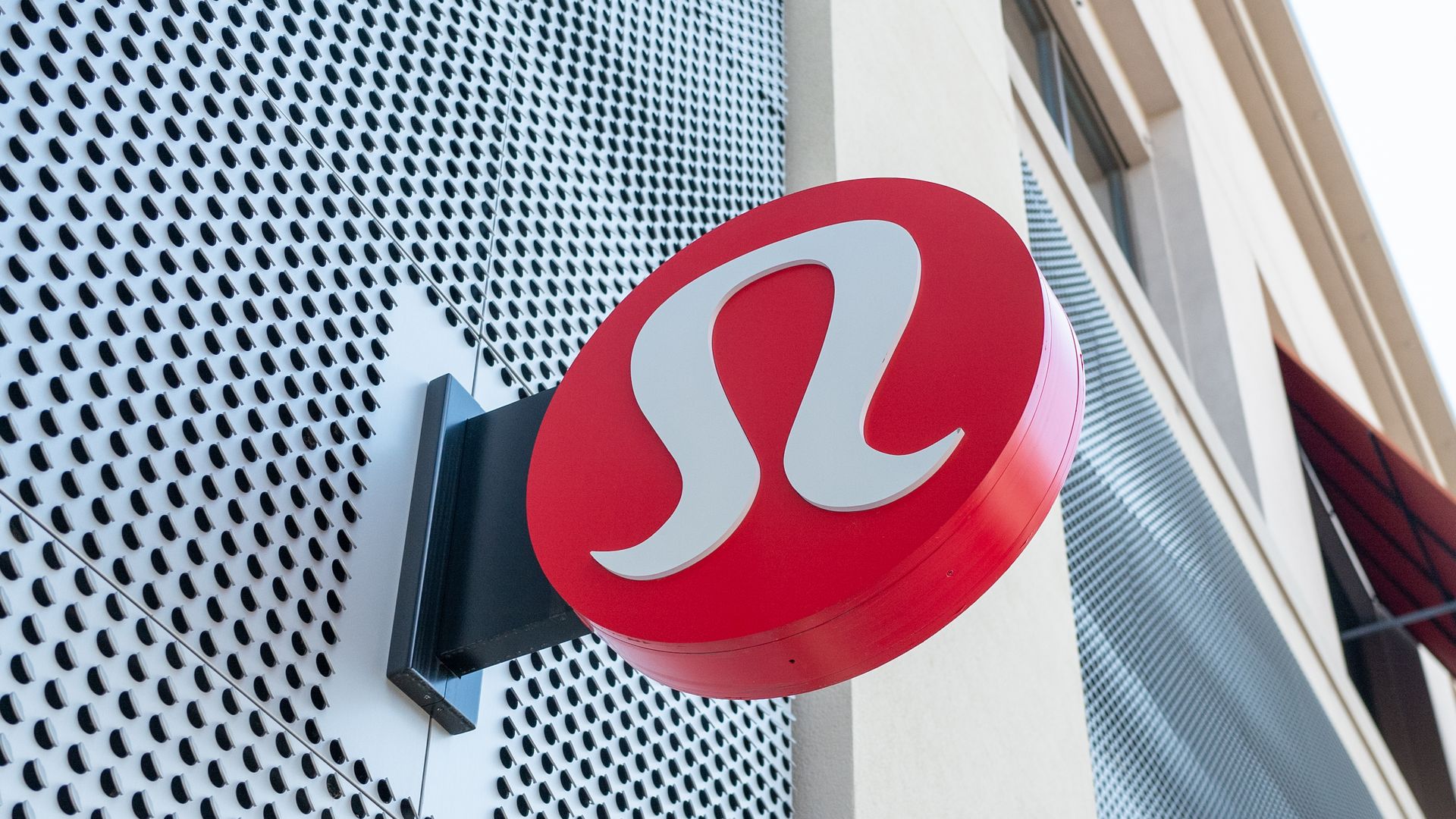 Red circular storefront sign with a white curved logo mounted on a beige building; a perforated metal wall fills the left background.