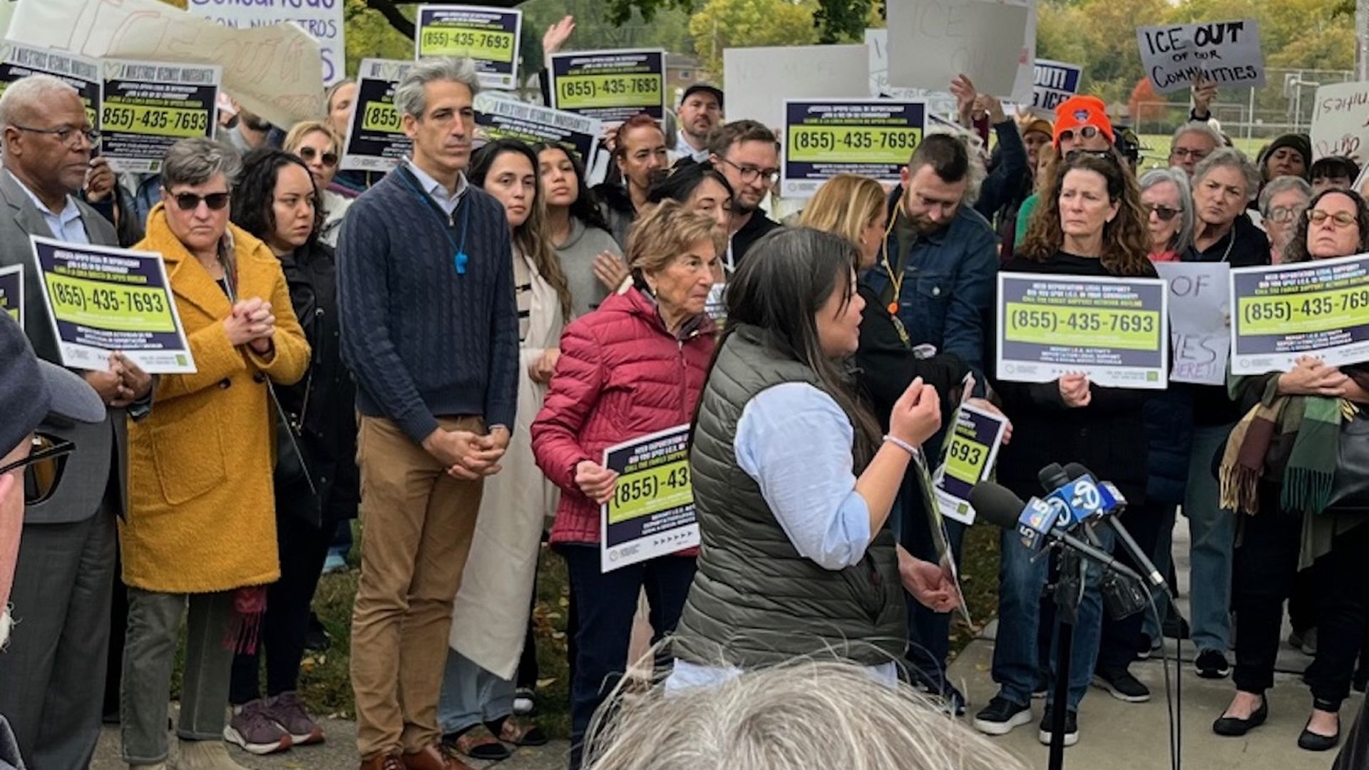 A diverse crowd holding signs with phone number (855)-435-7693 and messages like "Solidaridad" and "ICE out of our communities" listens to a speaker at a rally outdoors in fall.