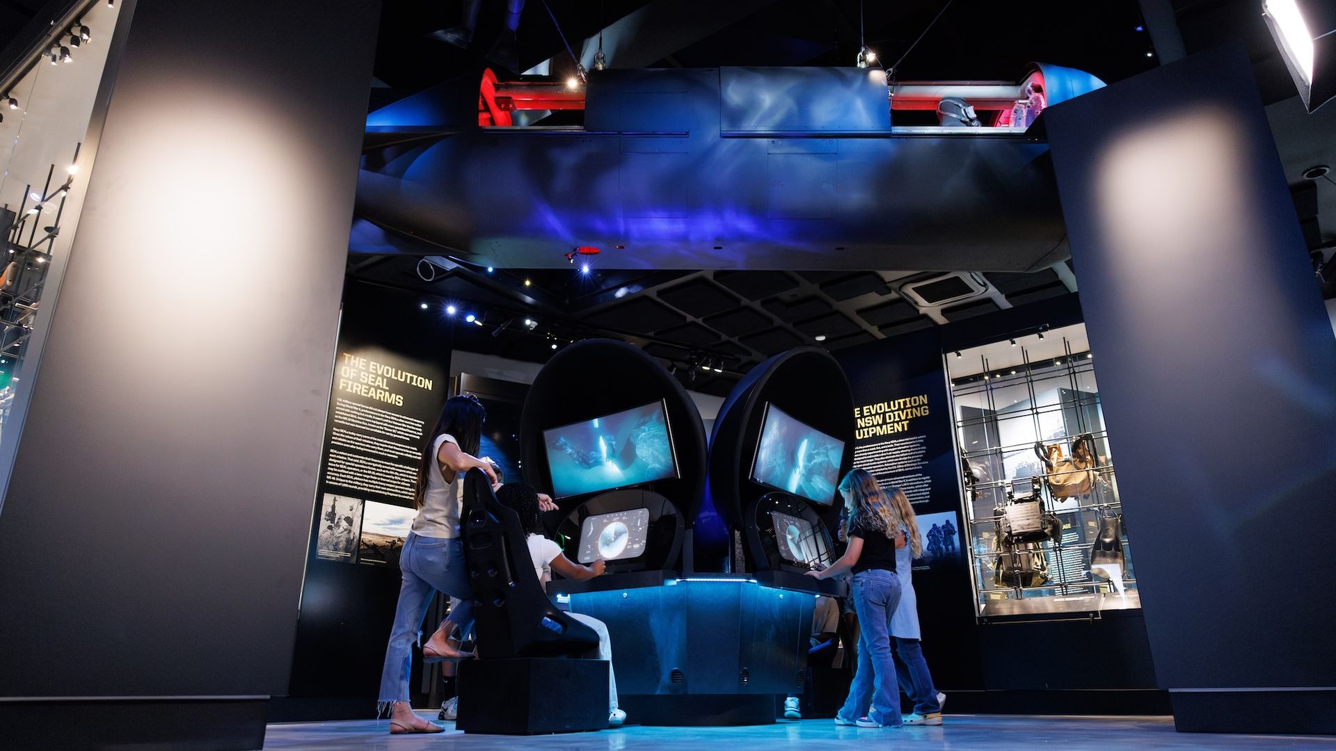 People interacting with two high-tech simulator displays under a suspended dark submersible in a dimly lit museum exhibit about Navy SEALs.