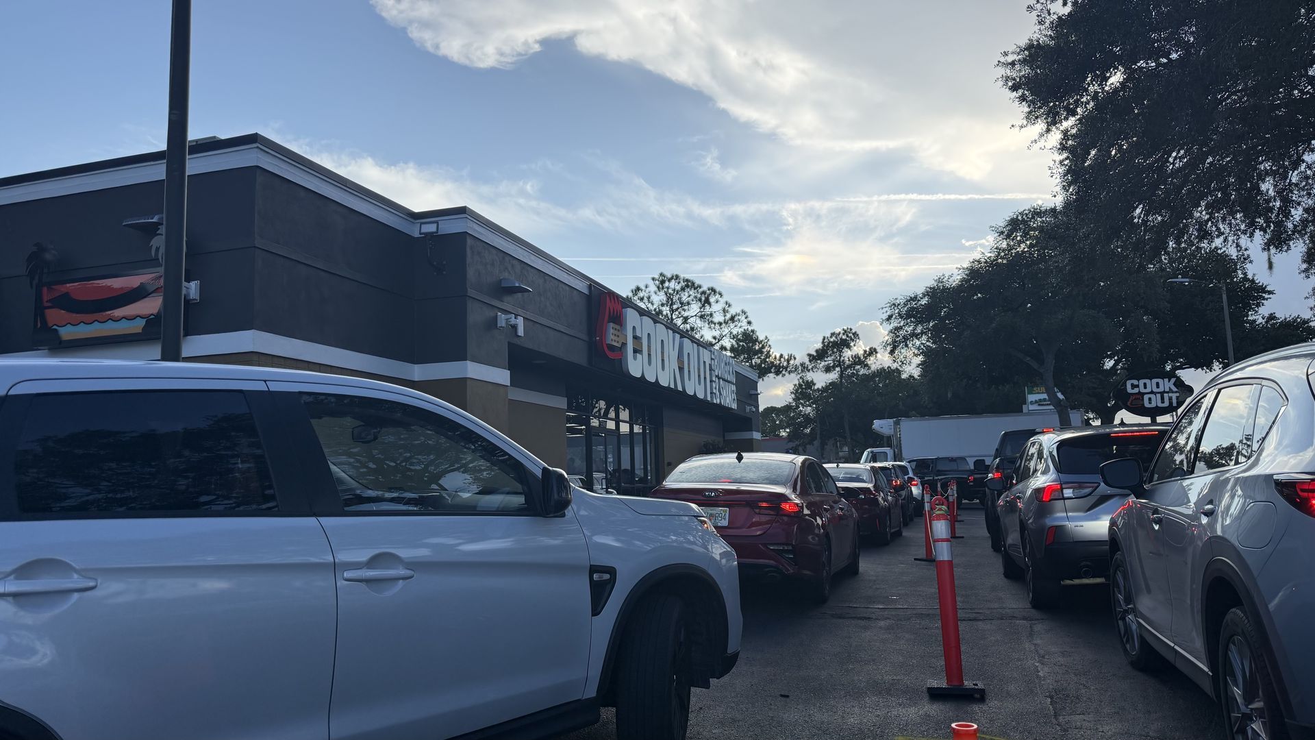 Cars line up at a Cook Out drive-thru; gray building with a large "Cook Out" sign, white SUV in the foreground, orange cones, trees on the right, and a blue sky with clouds.