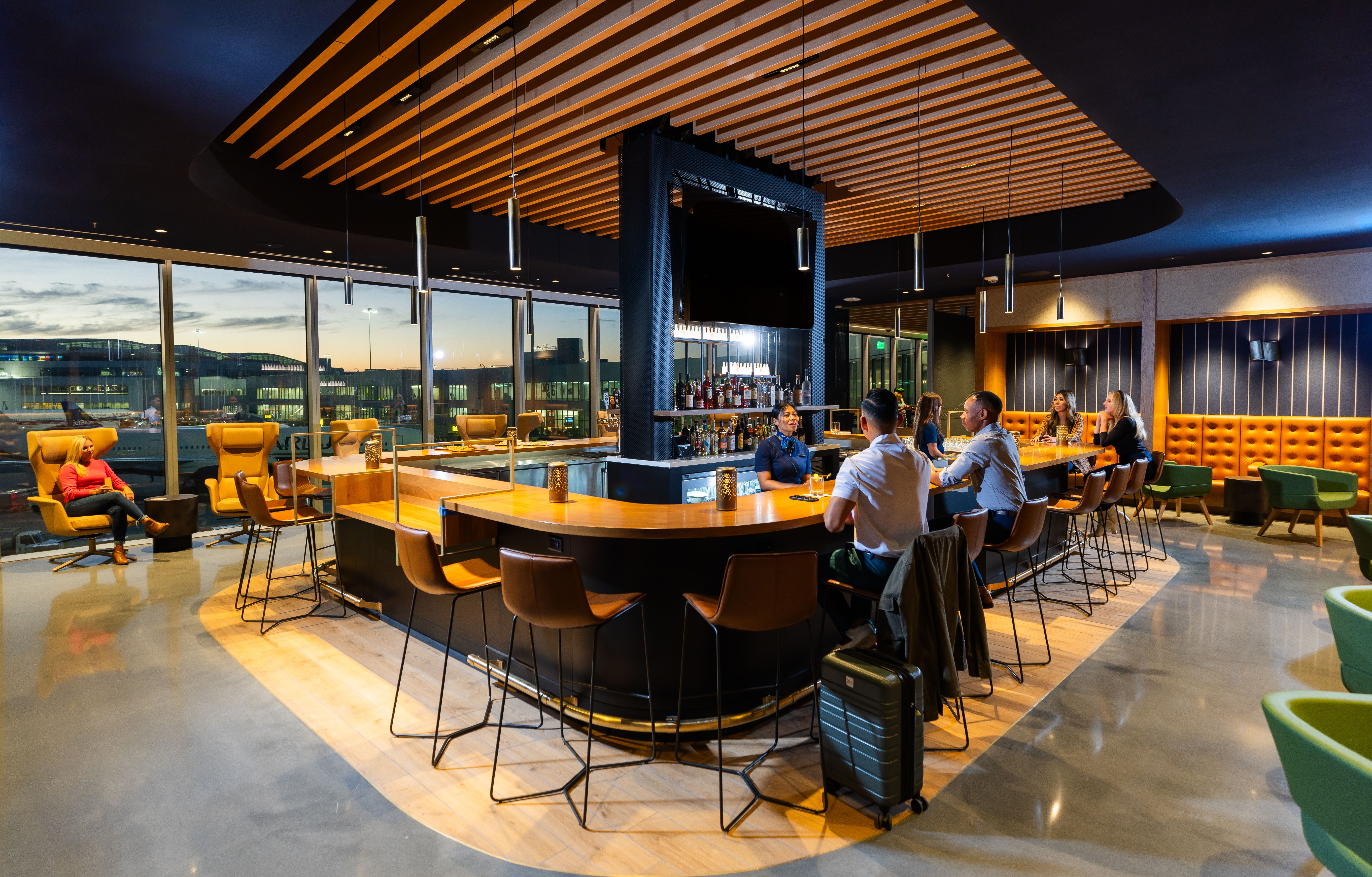 Modern airport lounge bar at sunset with patrons sitting on brown stools around a wooden bar, large windows showing planes outside, and orange and green seating along walls.