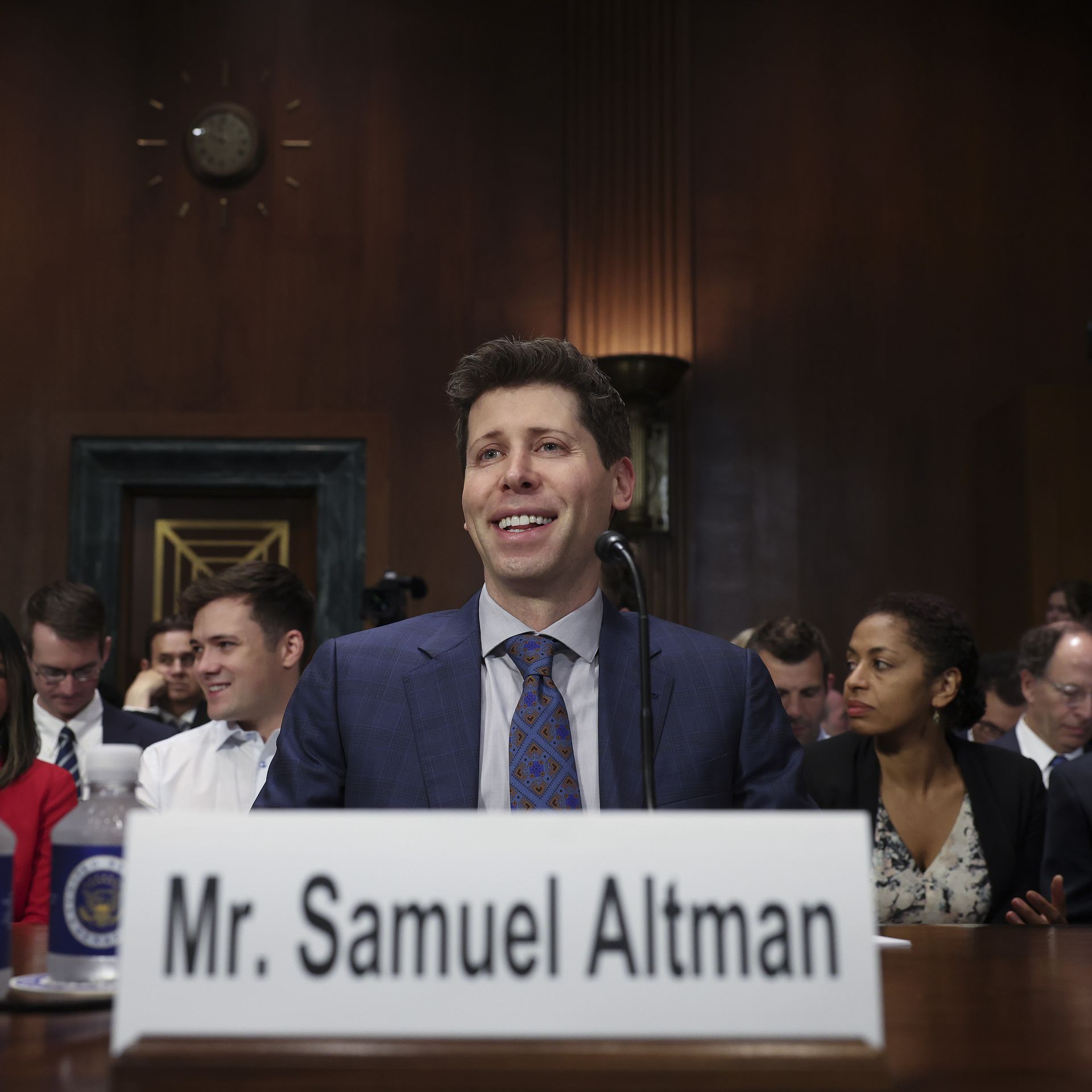 Photo of Sam Altman in suit and tie smiling, seated at testimony table, with a placard bearing his name