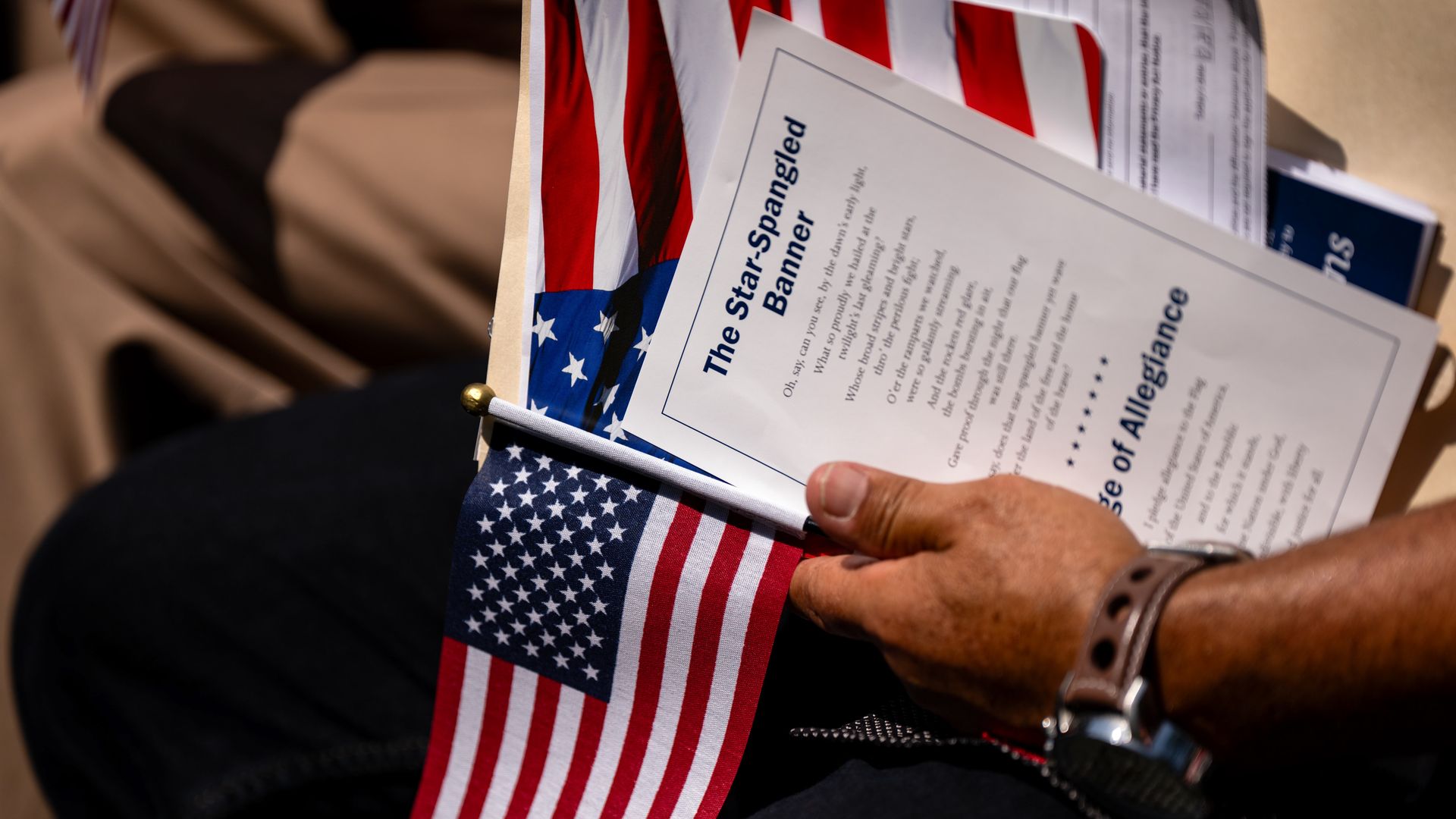 An attendee wearing a watch holds a small US flag and the lyrics of the Star Spangled Banner during a naturalization ceremony.