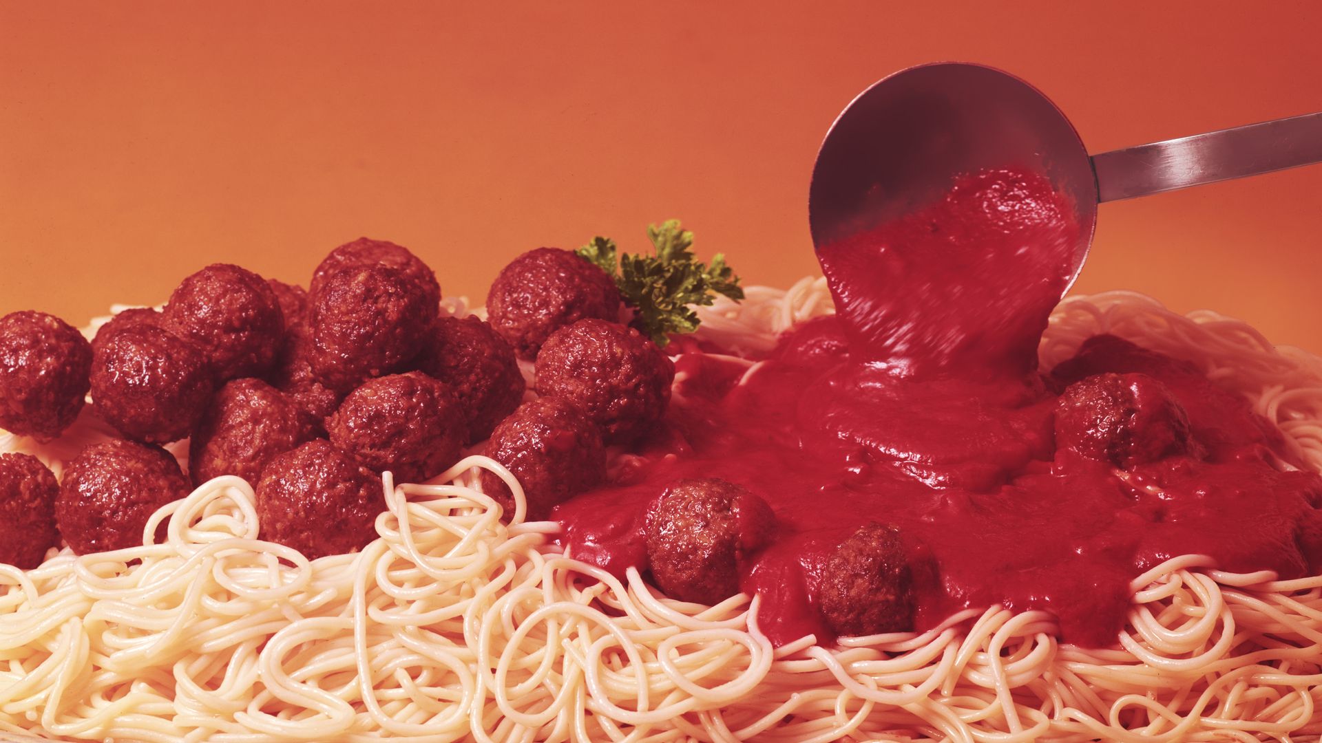 Pasta and meatballs on an orange background, with a silver ladle spreading spaghetti sauce.