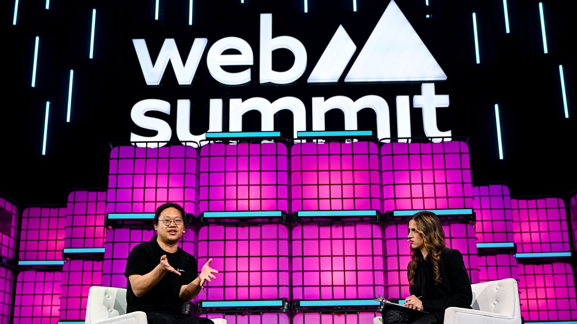 Two panelists sit on white chairs on a stage with a large "Web Summit" sign and mountain logo; a pink grid backdrop and blue neon strips frame the scene.