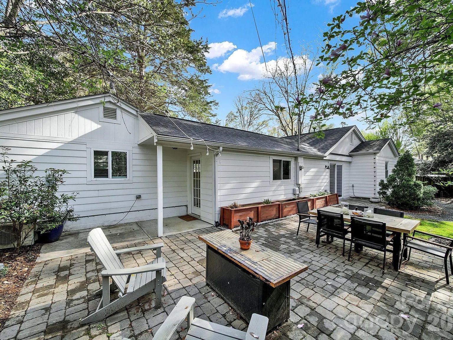 Outdoor patio area with white Adirondack chairs, a wooden table with potted plant, and dining table with black chairs, set against a white house with trees and blue sky overhead.