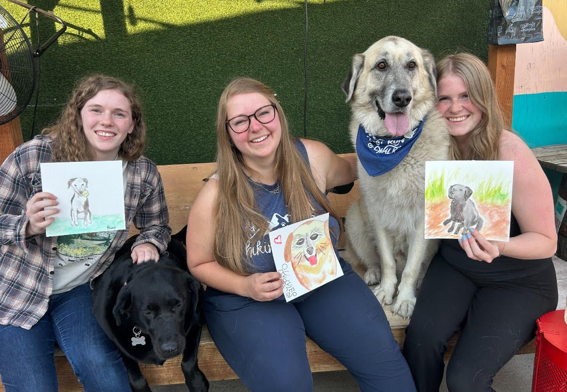 Three women sit holding water color pictures of dogs, with a brown and tan colored dog sitting between two of them and a black dog lying down next to another.