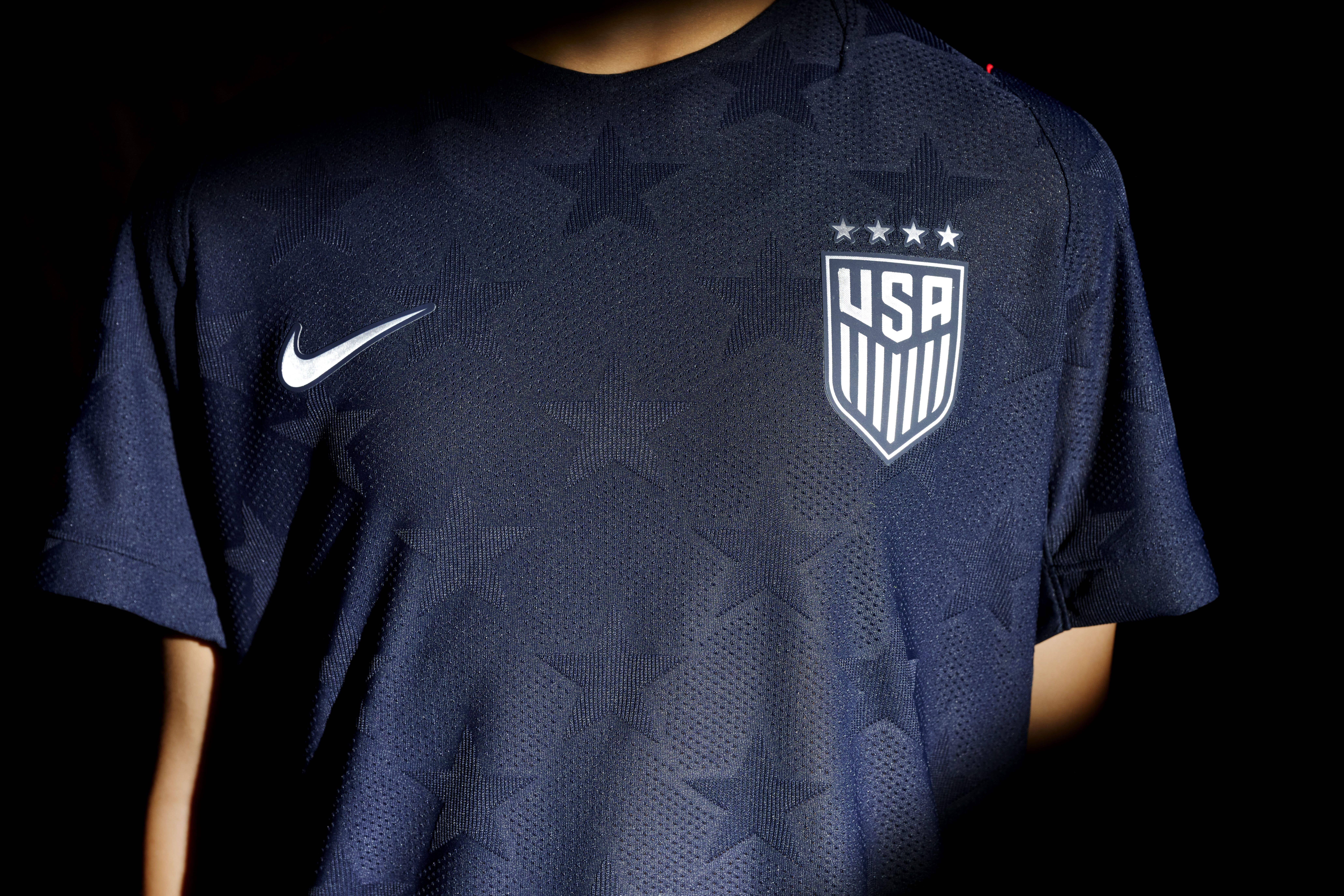 Dark blue USA soccer jersey with a white Nike swoosh on the left and the USA crest with five stars above on the right; the fabric shows a subtle star pattern.