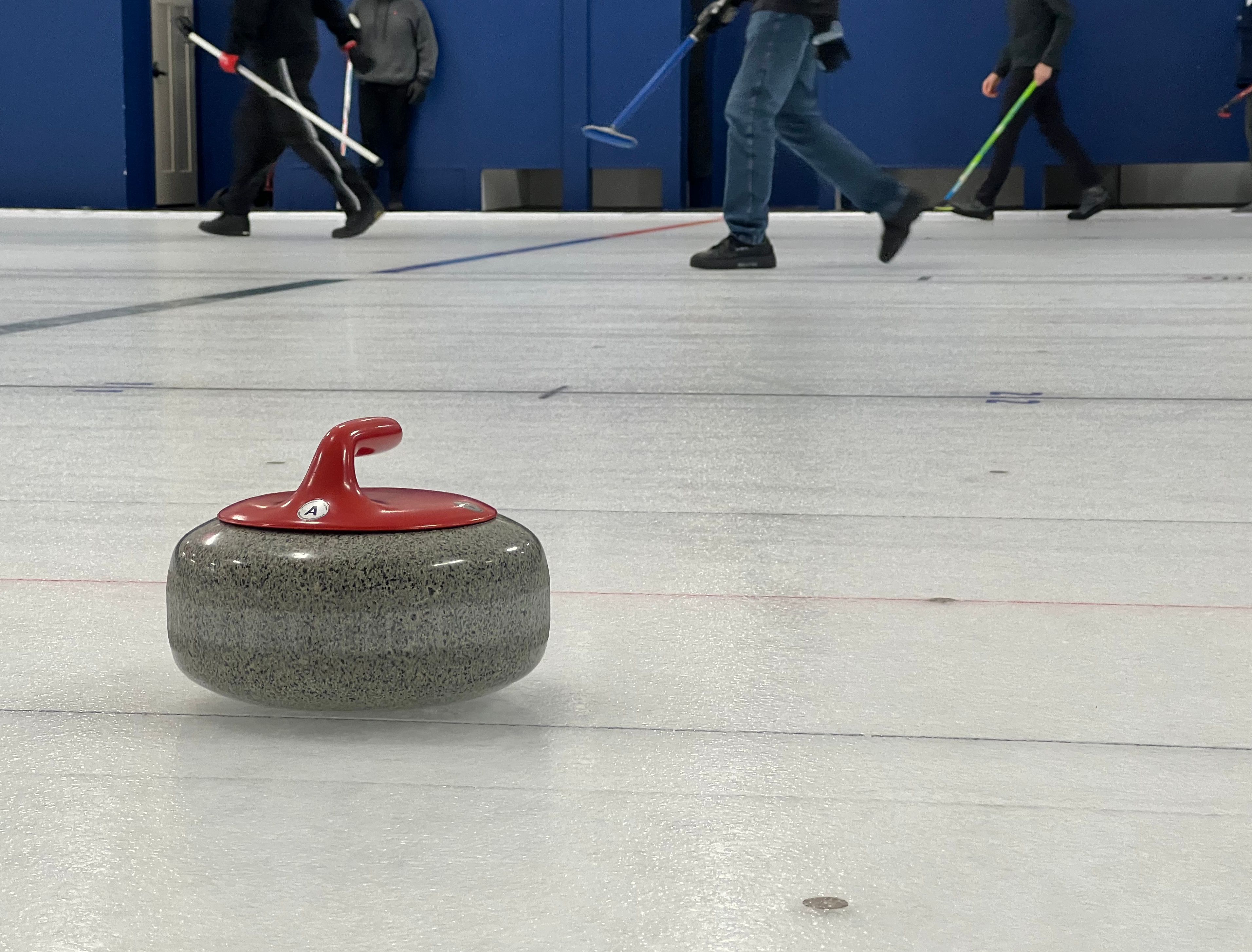 A curling stone with a red handle sits on a white sheet of ice with players' feet in the background.