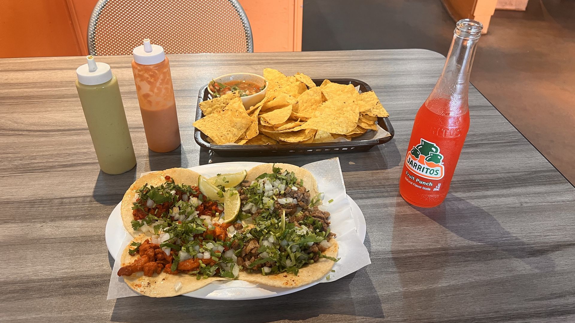 Table with three tacos topped with onions, cilantro, and lime wedges; basket of tortilla chips with salsa; two sauce bottles; and a bottle of Jarritos fruit punch soda.