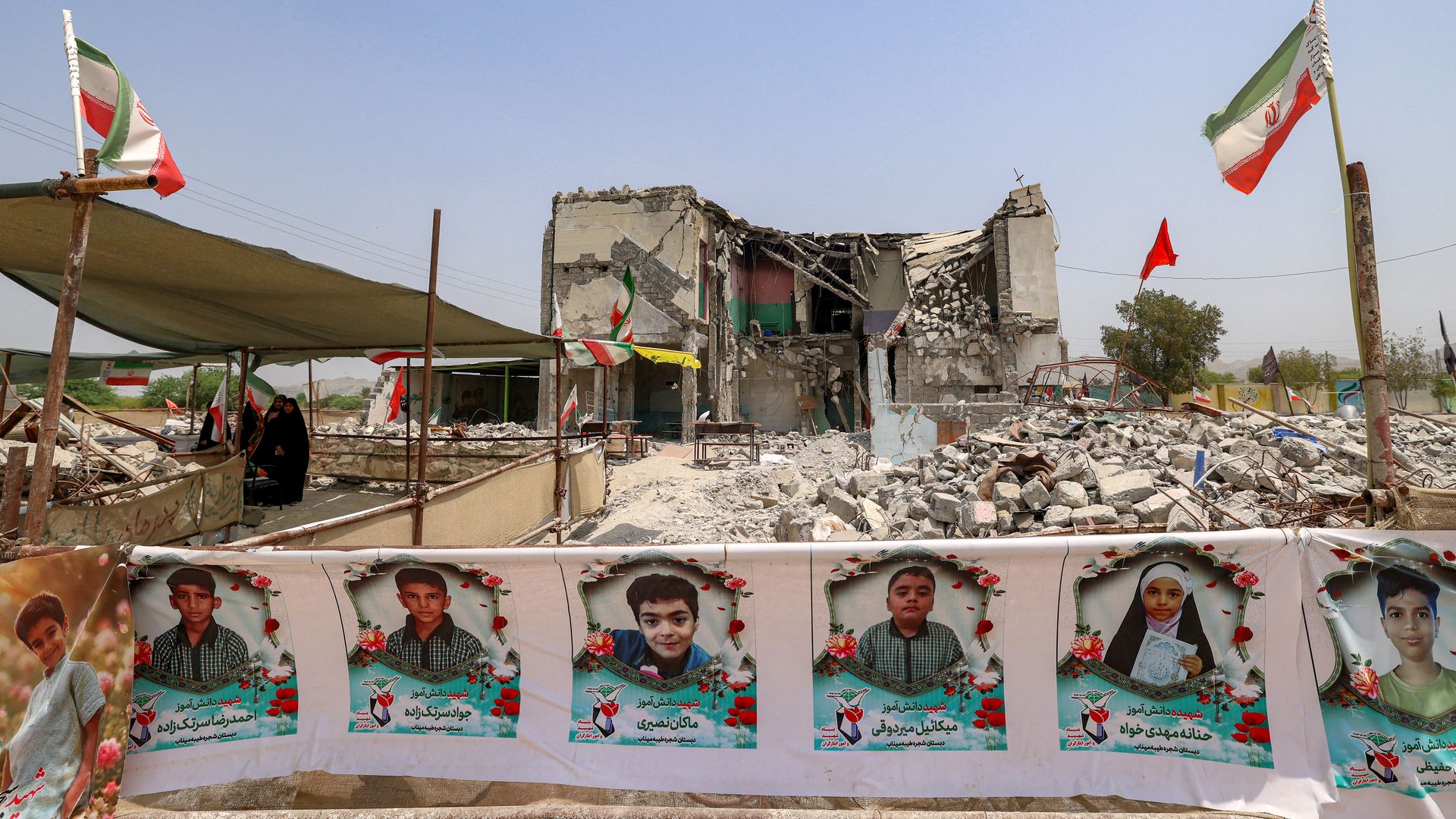 The building of a destroyed primary school is pictured surrounded by rubble. Iranian flags fly nearby and a wall displays pictures of several children.