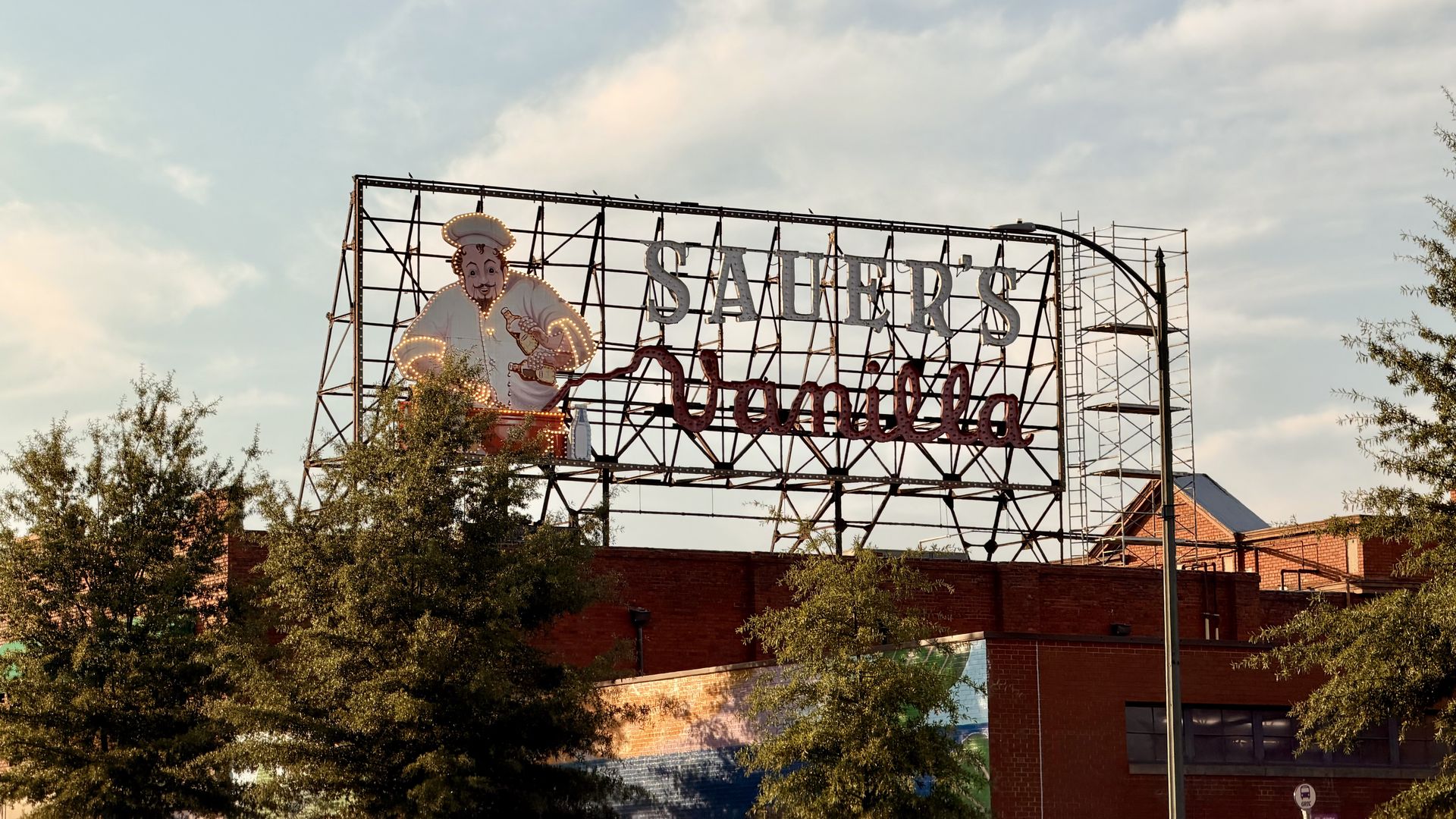 Large open metal sign frame with illuminated letters reading "SAUER'S" and a cartoon chef, mounted above a brick building; trees in the foreground and a blue sky.