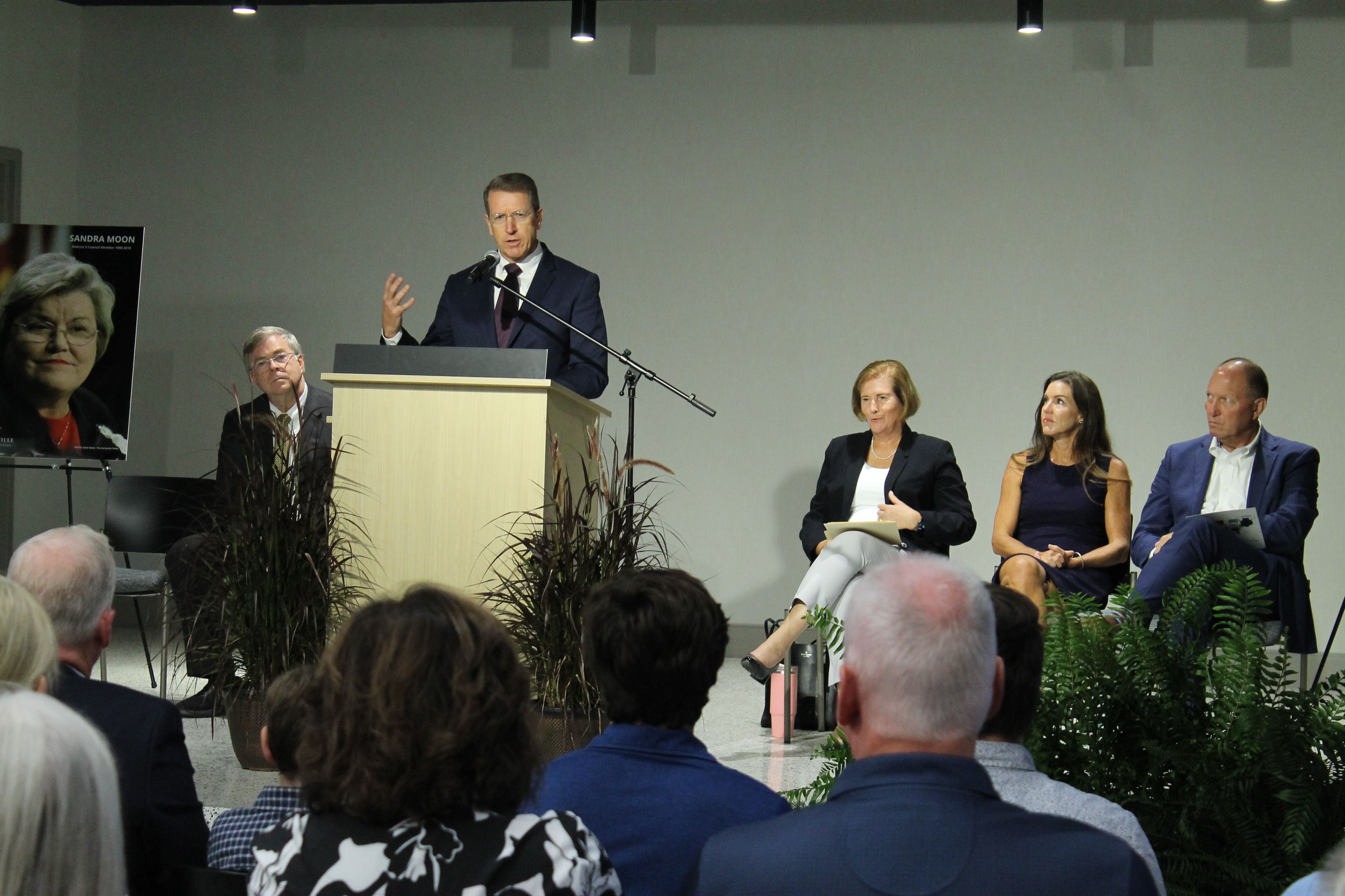 Man in a dark suit and maroon tie speaks at a wooden podium on stage, microphone extended; four panelists sit right. A poster of a woman is left; audience and plants in foreground.