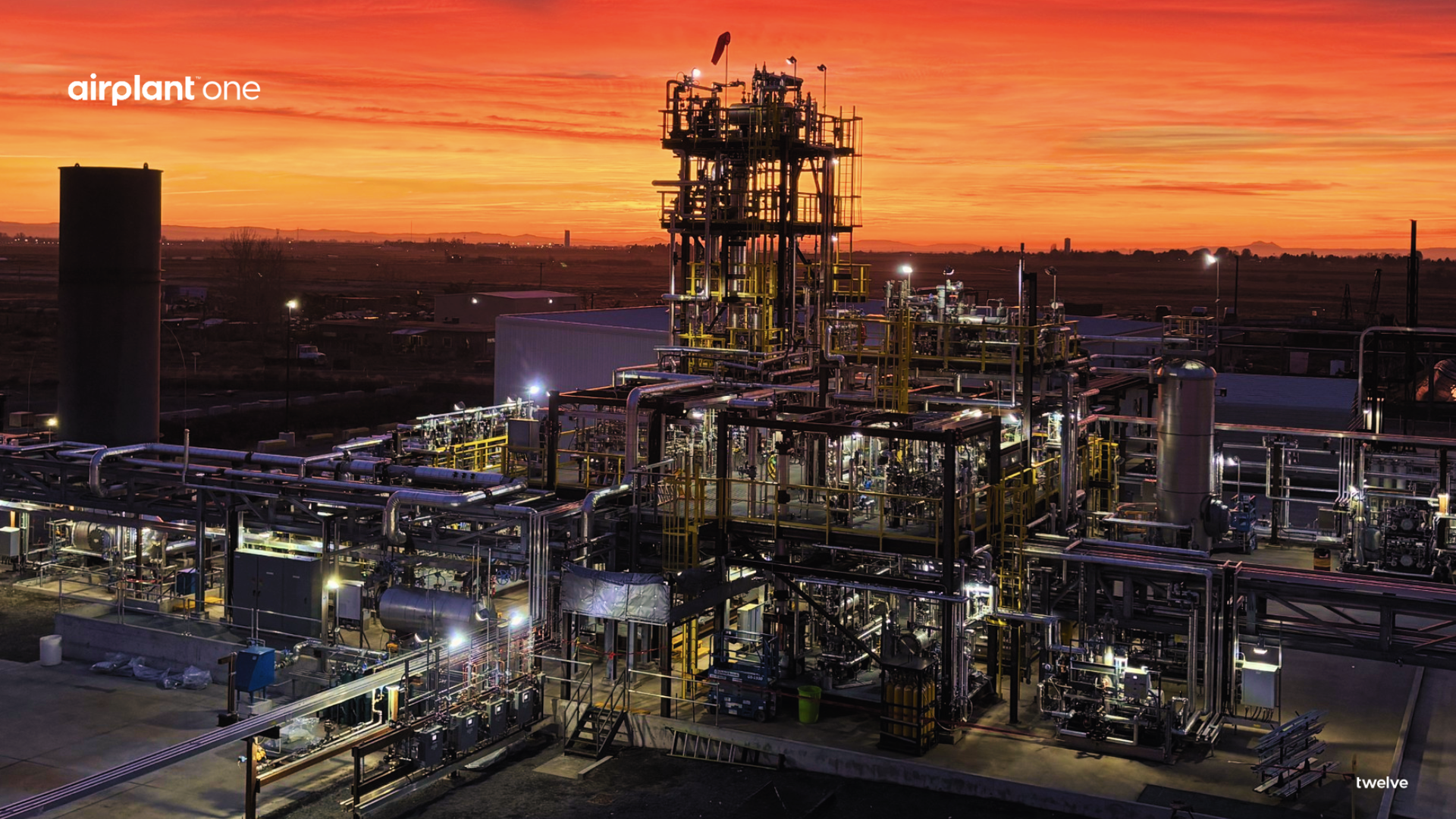 Industrial refinery at sunset with a dense network of pipes and catwalks, a tall processing tower at center-right, bright lights, and a fiery orange horizon; logo airplant one on the left.