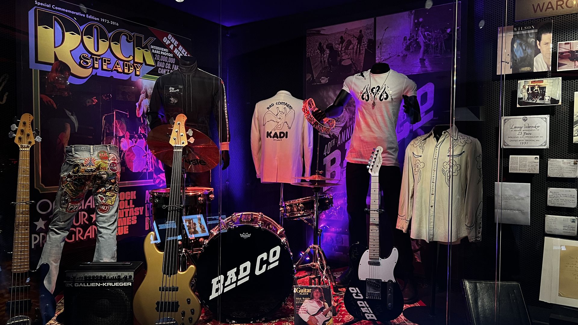 Display of Bad Company band memorabilia including guitars, bass, drum set, vintage jackets, shirts, and music-related posters under purple and red lighting.