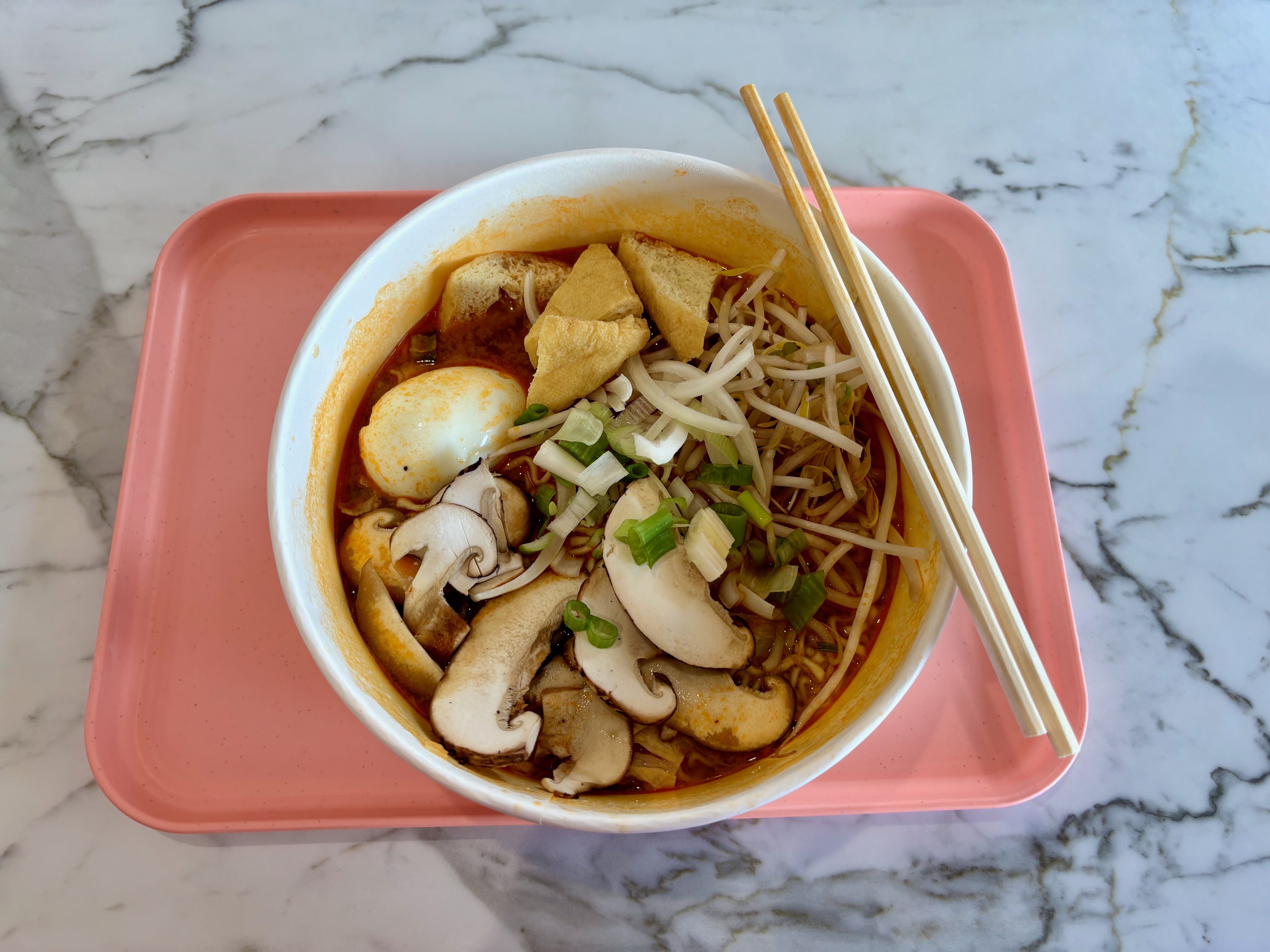 Top-down view of a white bowl of ramen on a pink tray, filled with orange-red broth, bean sprouts, green onions, sliced mushrooms, a soft-boiled egg, and tofu skins. Chopsticks rest on the bowl.