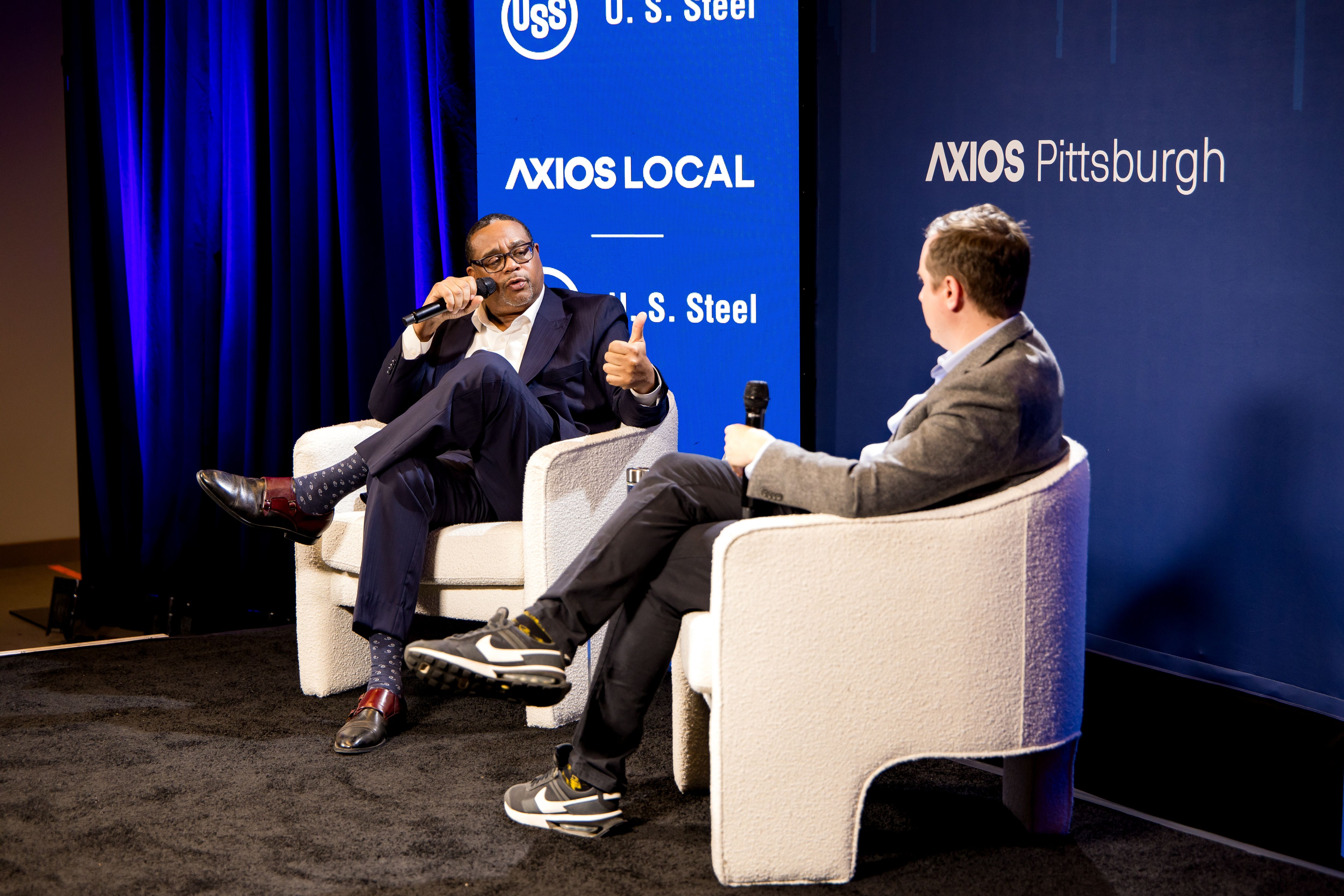 Two men sit in white chairs on a stage in front of a blue backdrop that says Axios Pittsburgh. They're both holding microphones in conversation with each other.