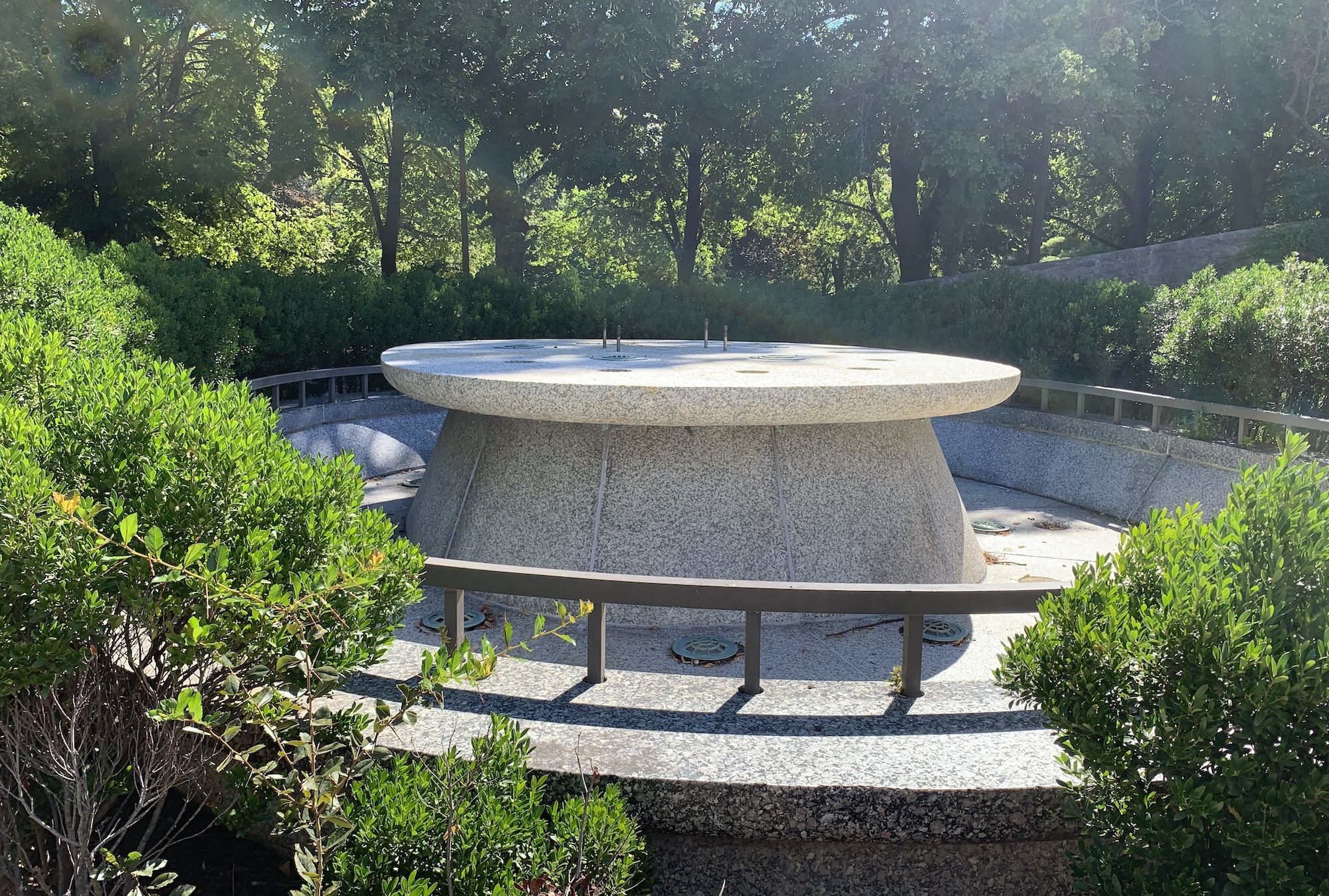 A large gray stone sundial surrounded by green bushes with trees and sunlight in the background in an outdoor garden setting.