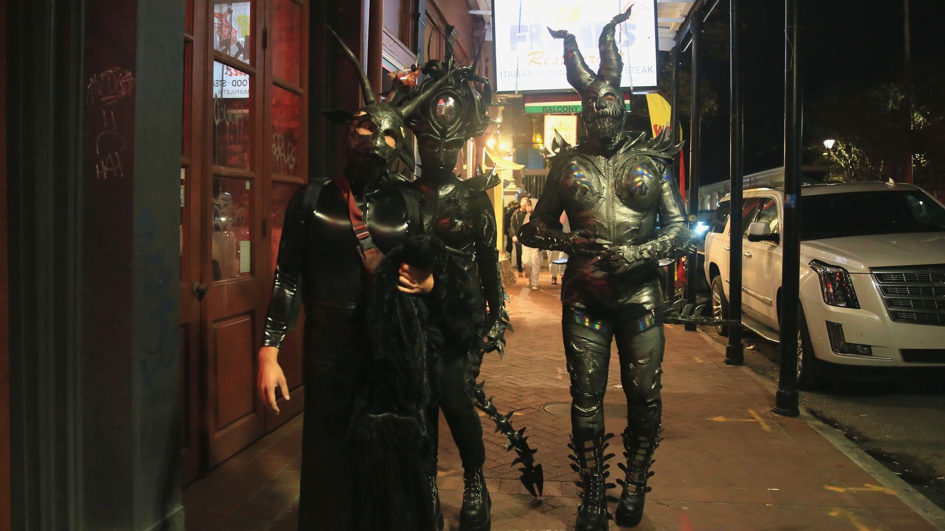 Three people in medieval-looking armor costumes walk in the French Quarter under a lighted store sign.