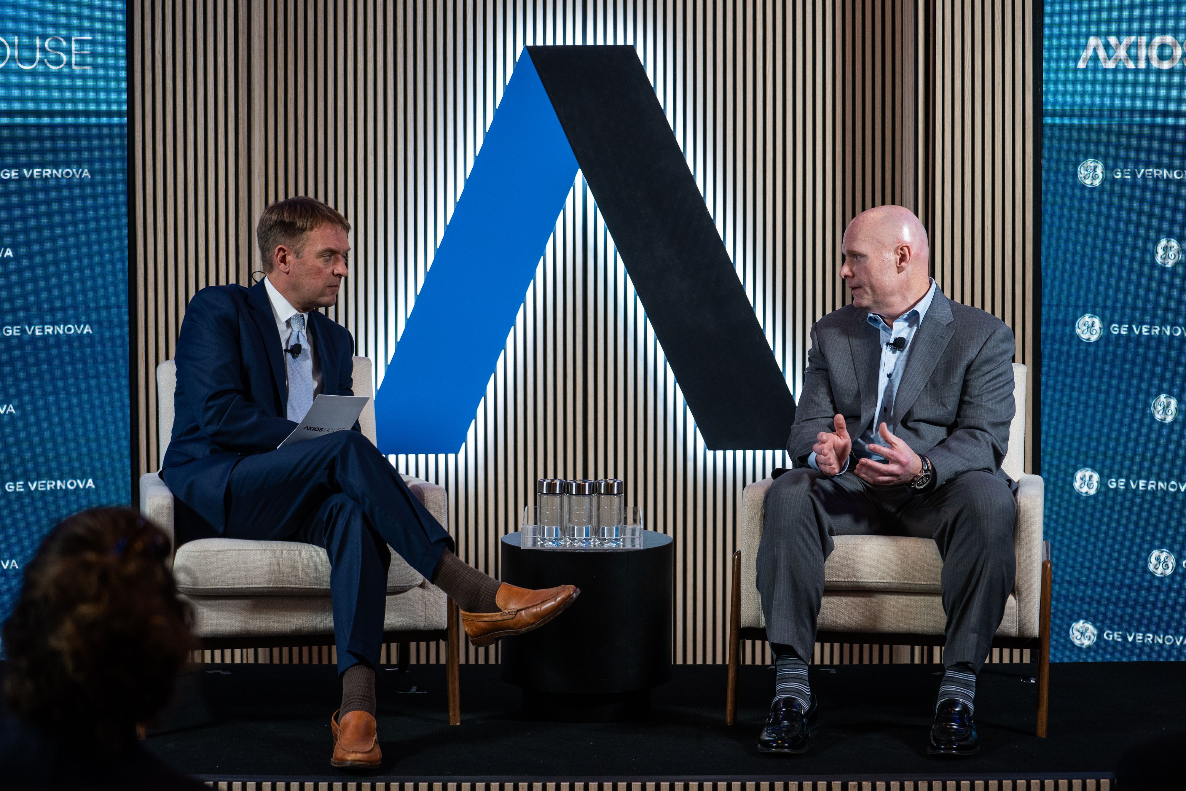 Two men in suits sit on a stage, wearing lapel mics. A large blue and black diagonal backdrop rises between them, flanked by GE Vernova banners; a glass table with water glasses sits between.