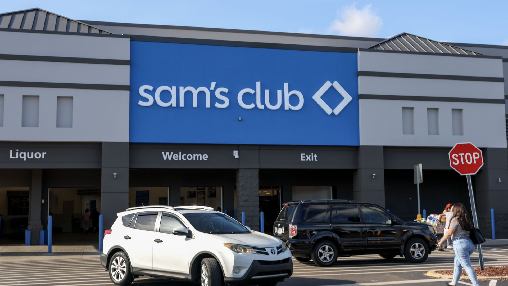 Exterior of a Sam's Club storefront with a large blue sign and white diamond logo. In front, a white SUV and a black SUV sit in the parking lot as a shopper walks past a red stop sign on a sunny day.