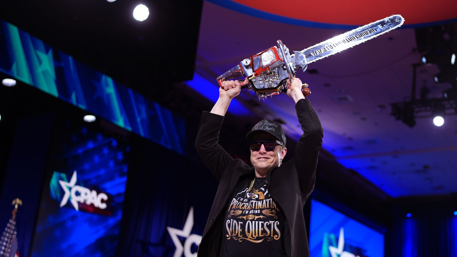 Elon Musk waves a chainsaw over his head while wearing a black MAGA hat and sunglasses at the Conservative Political Action Conference.