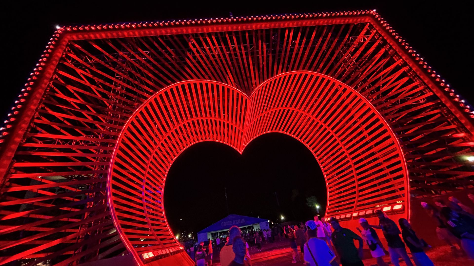 Night scene of a large red illuminated arch sculpture with radial spokes forming a heart-shaped opening; red lights line the frame while a crowd gathers beneath against a dark sky.