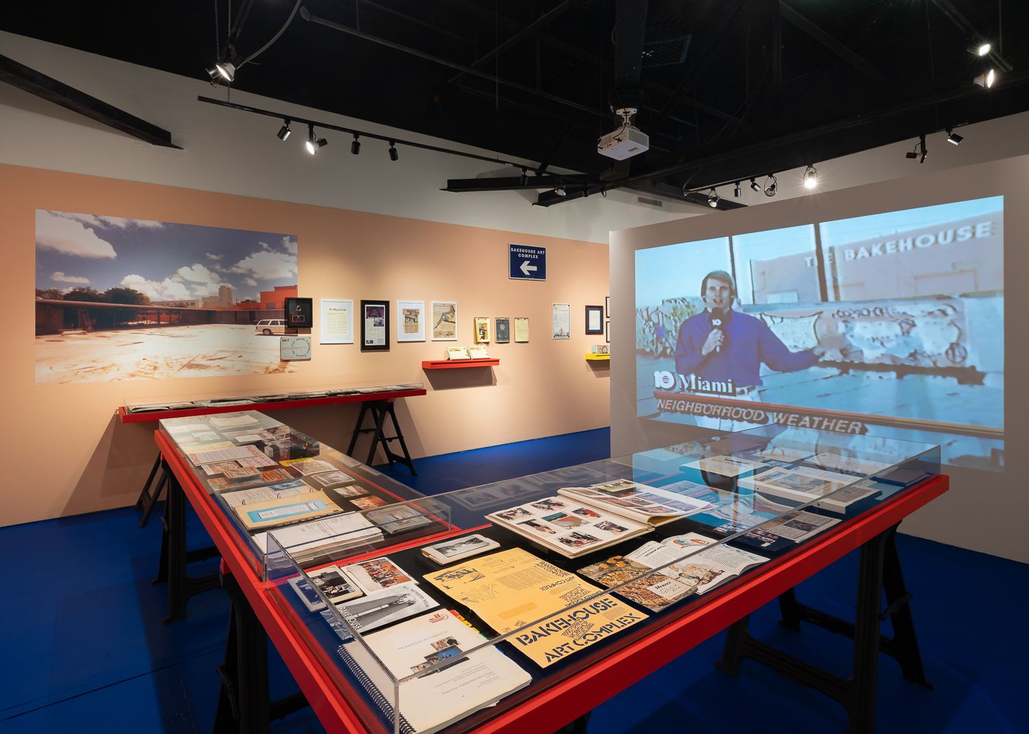 Exhibit in a gallery with a blue floor, red display tables with documents under glass, a large photo on the wall, and a projected video of a reporter in front of The Bakehouse.