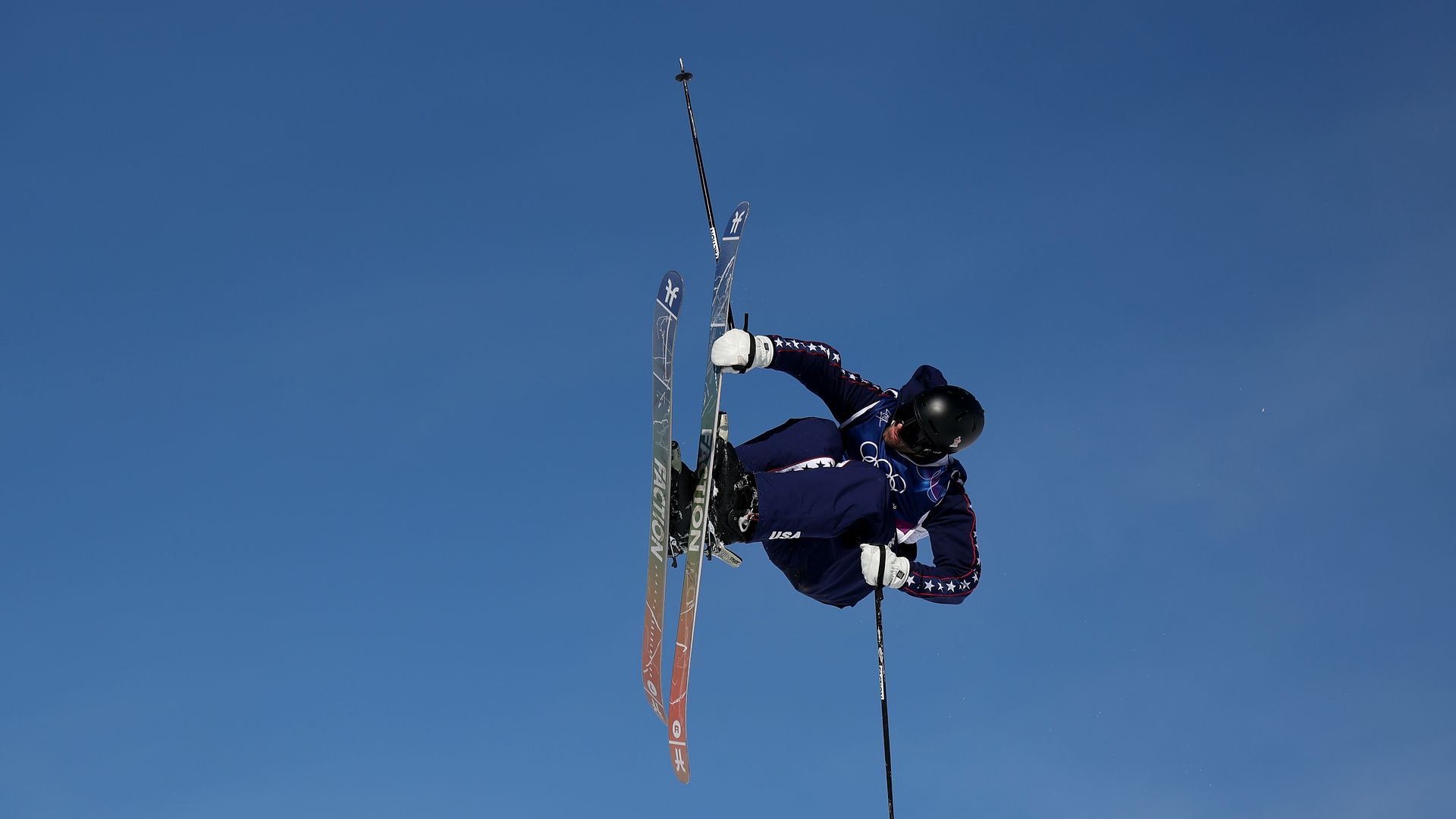 A skier in a black uniform soars into the air with a blue sky behind him.