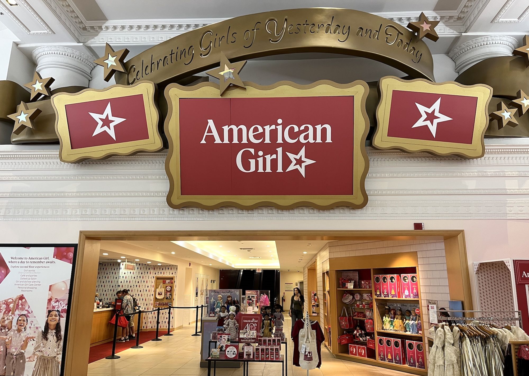 Storefront for "American Girl" with red and gold signage and star motifs; curved banner reads "Celebrating Girls of Yesterday and Today." Inside, bright shelves display dolls and pink products.