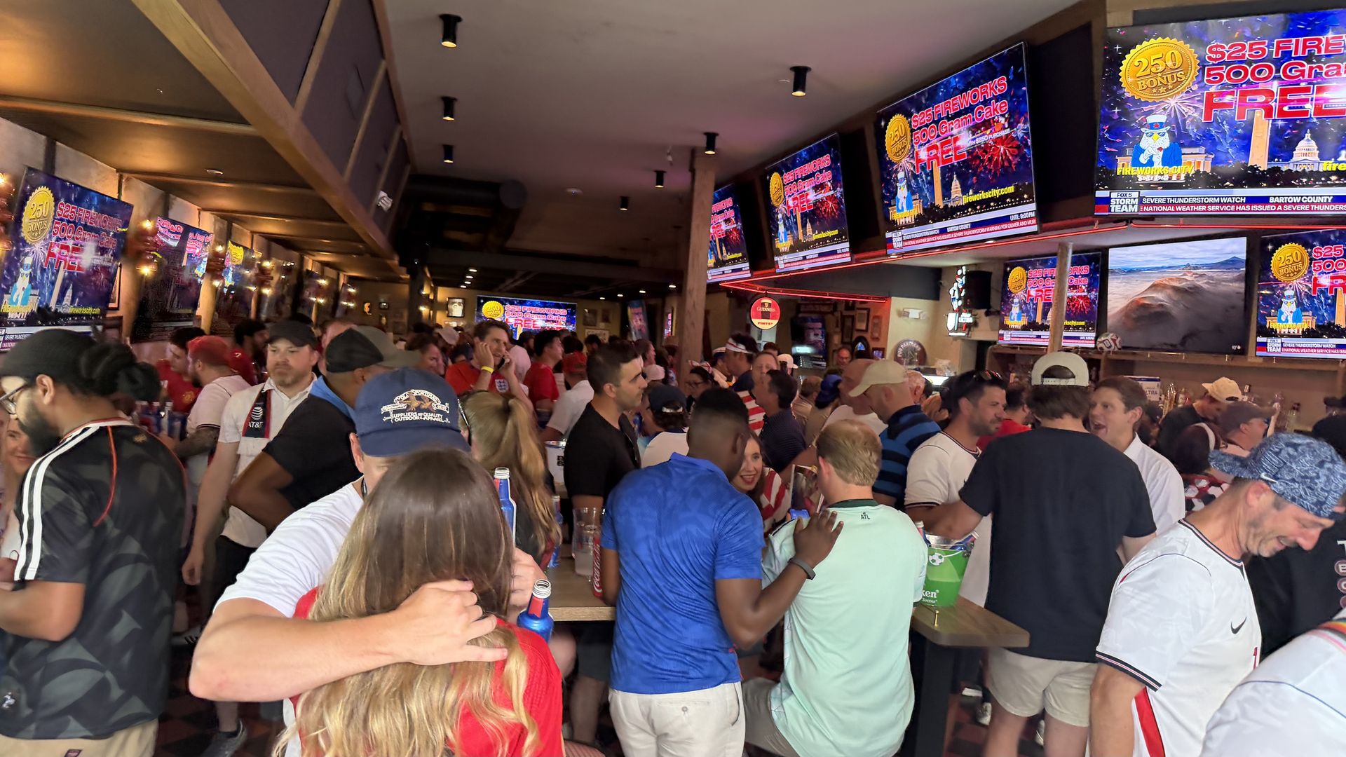 Crowded indoor sports bar with many patrons standing and chatting; wall-mounted TVs display bright fireworks promotions and logos, under warm lighting and wooden beams.