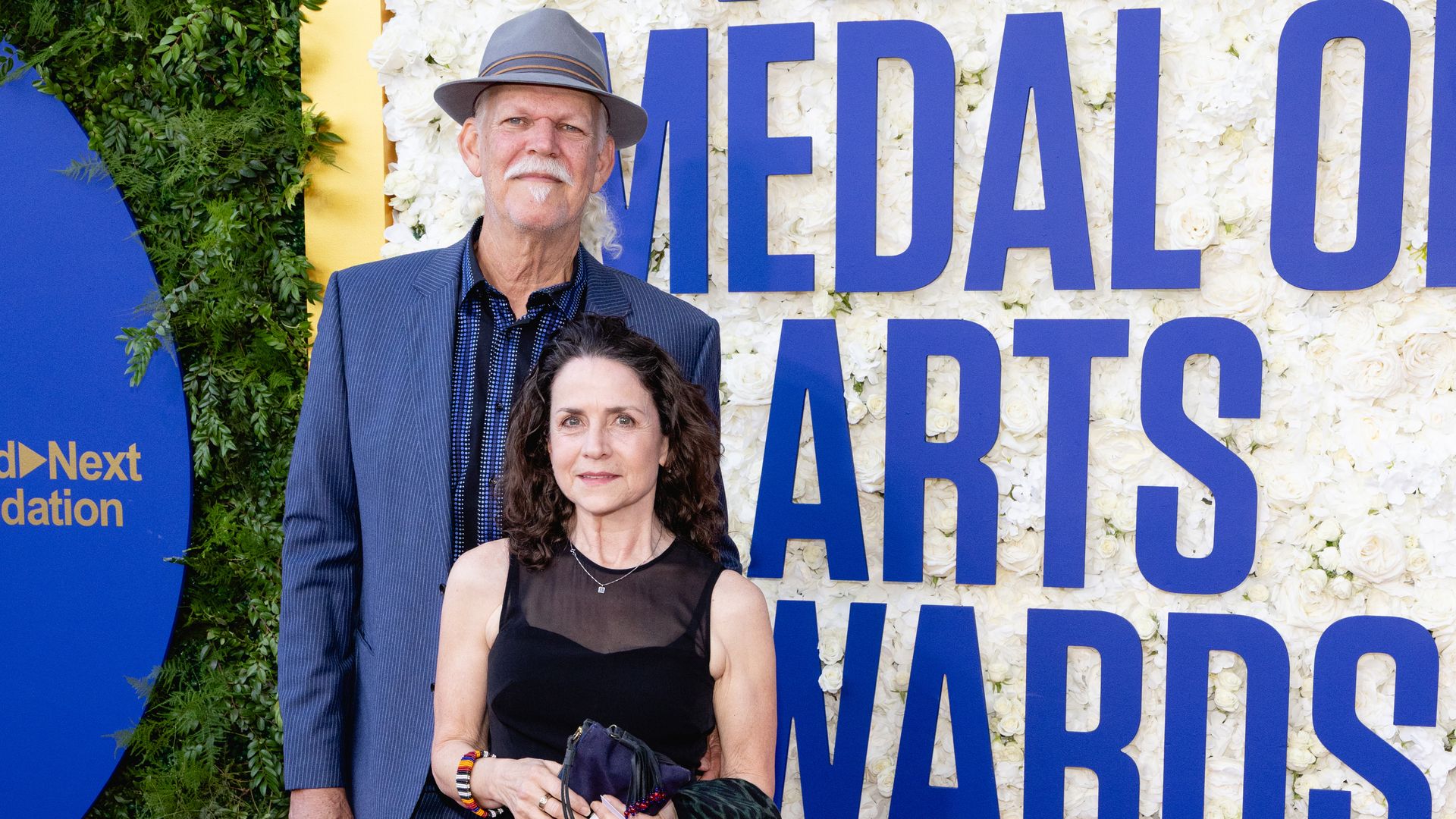 Two people pose in front of a white flower wall with large blue letters reading "Medal of Arts Awards". An older man wearing a gray fedora and pinstriped suit stands beside a woman in a black dress.