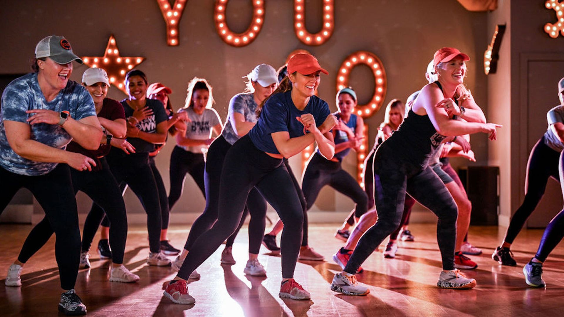 A group of women in athletic wear and caps, dancing energetically in a studio with wooden floors and illuminated letters spelling. 