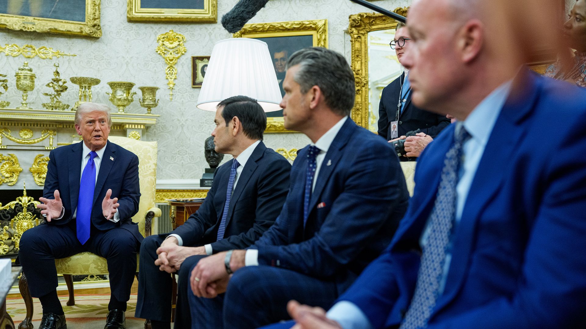 Trump, wearing a dark suit and a blue tie, gestures as he speaks in the Oval Office with. Several other men in dark suits sitting on a couch watch him as he speaks.