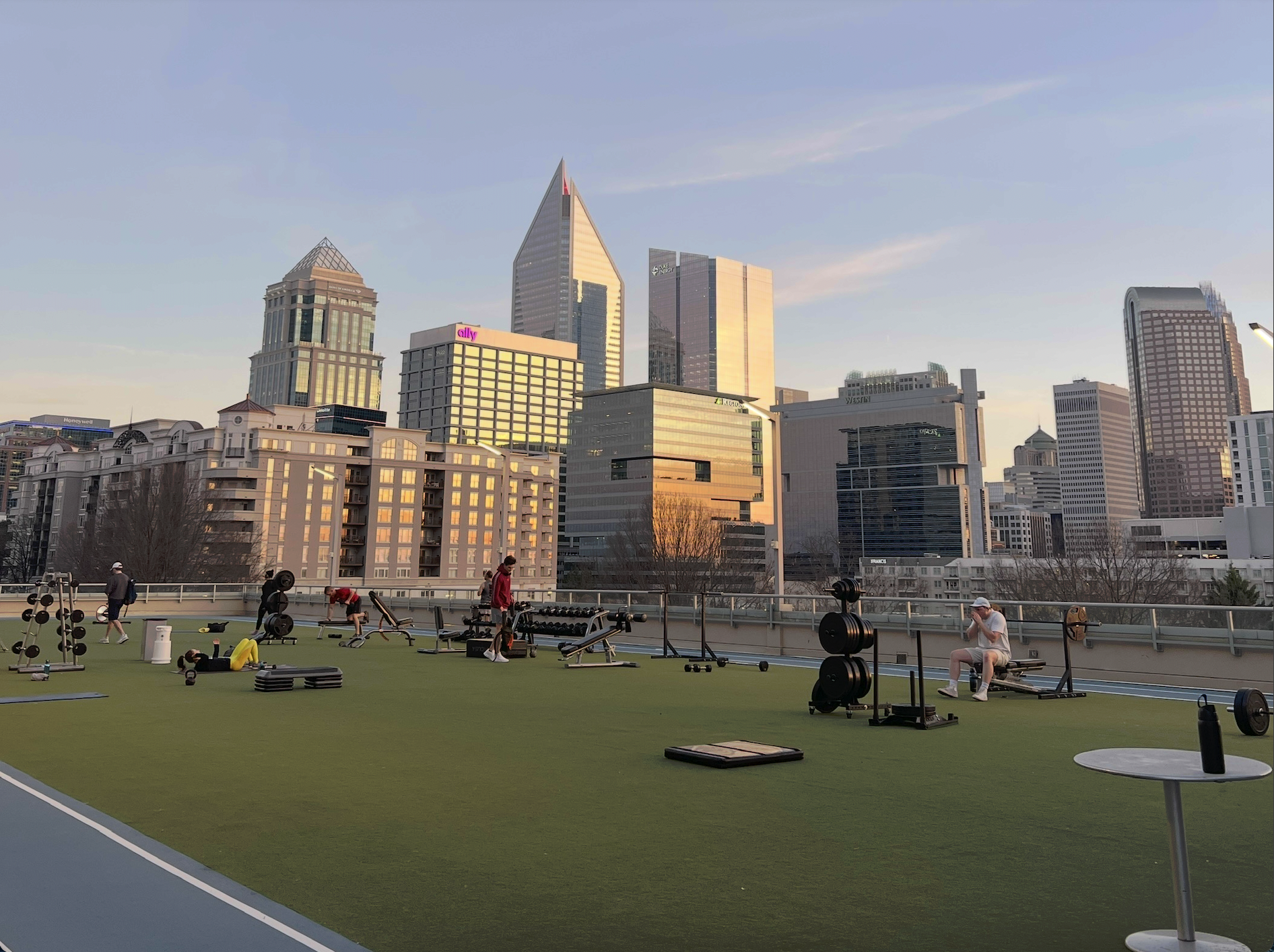 Rooftop gym on a city skyline: people lifting weights on green turf with benches and dumbbells, overlooking tall modern buildings bathed in sunset light.
