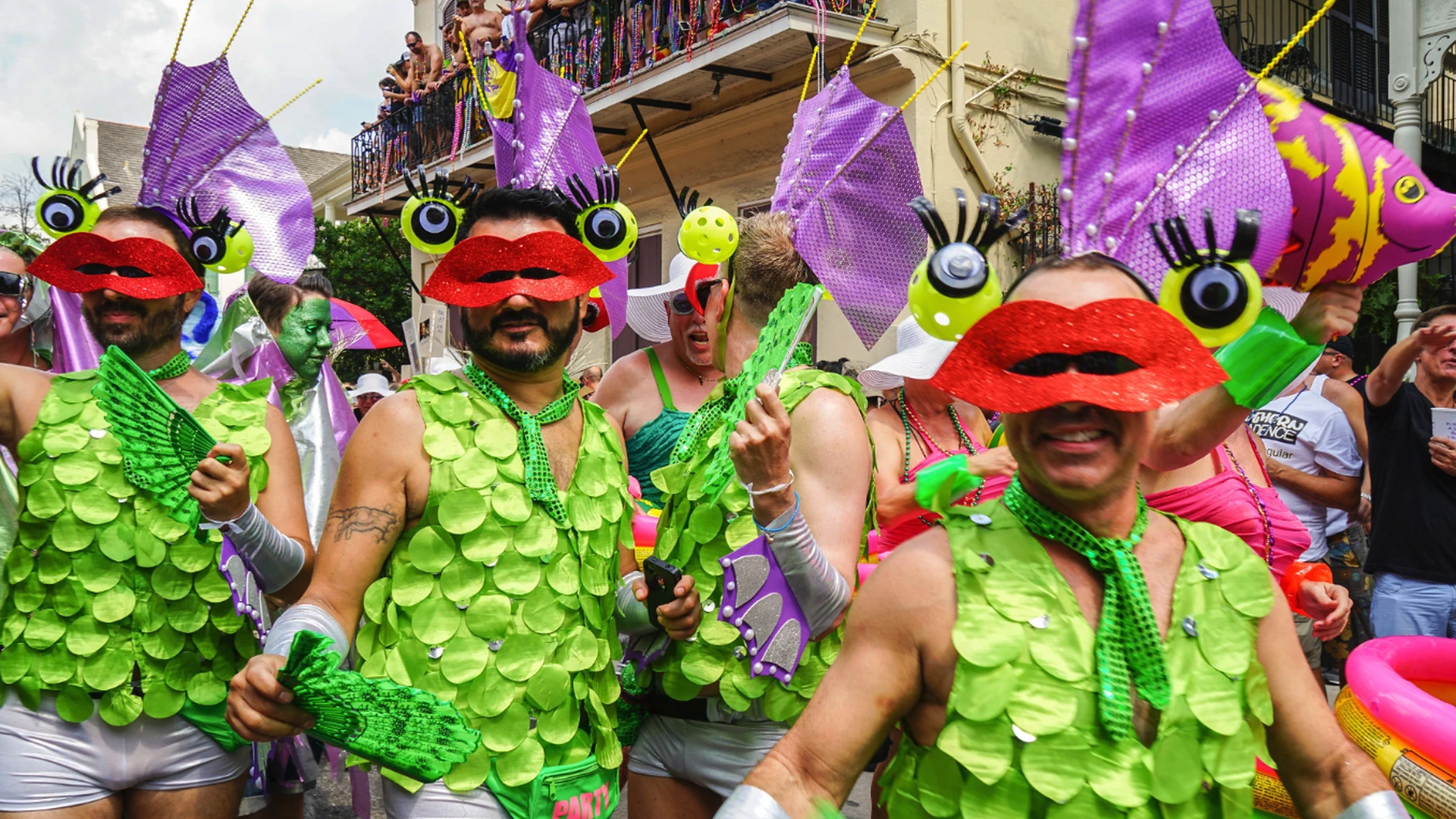 Vibrant street parade with participants in lime-green scale costumes and red glitter lips masks, oversized purple fish hats, neon green accessories, and a crowd watching from shopfronts.