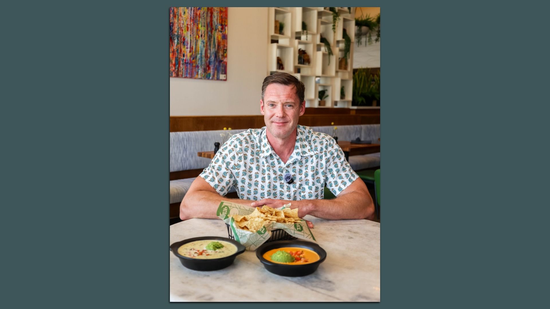 Smiling man in a pineapple-patterned shirt sits at a marble table in a bright restaurant, with bowls of chips and dips in front. Colorful abstract art on the wall and plant-filled shelves behind him.