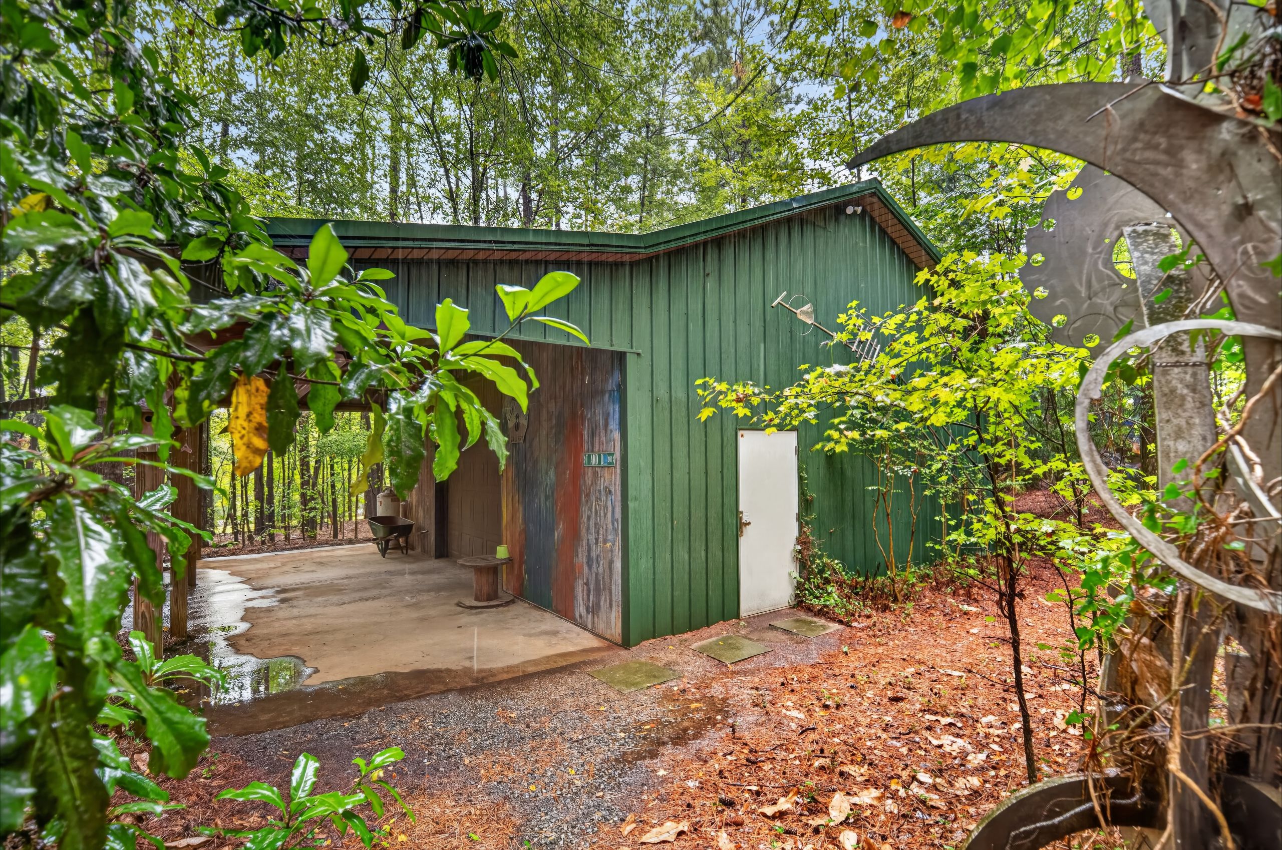 Green wooden shed with a sloped roof sits in a lush, rain-soaked garden. Dense trees and leaves frame the scene; a circular metal sculpture on the right, and a wet concrete path runs along the shed.